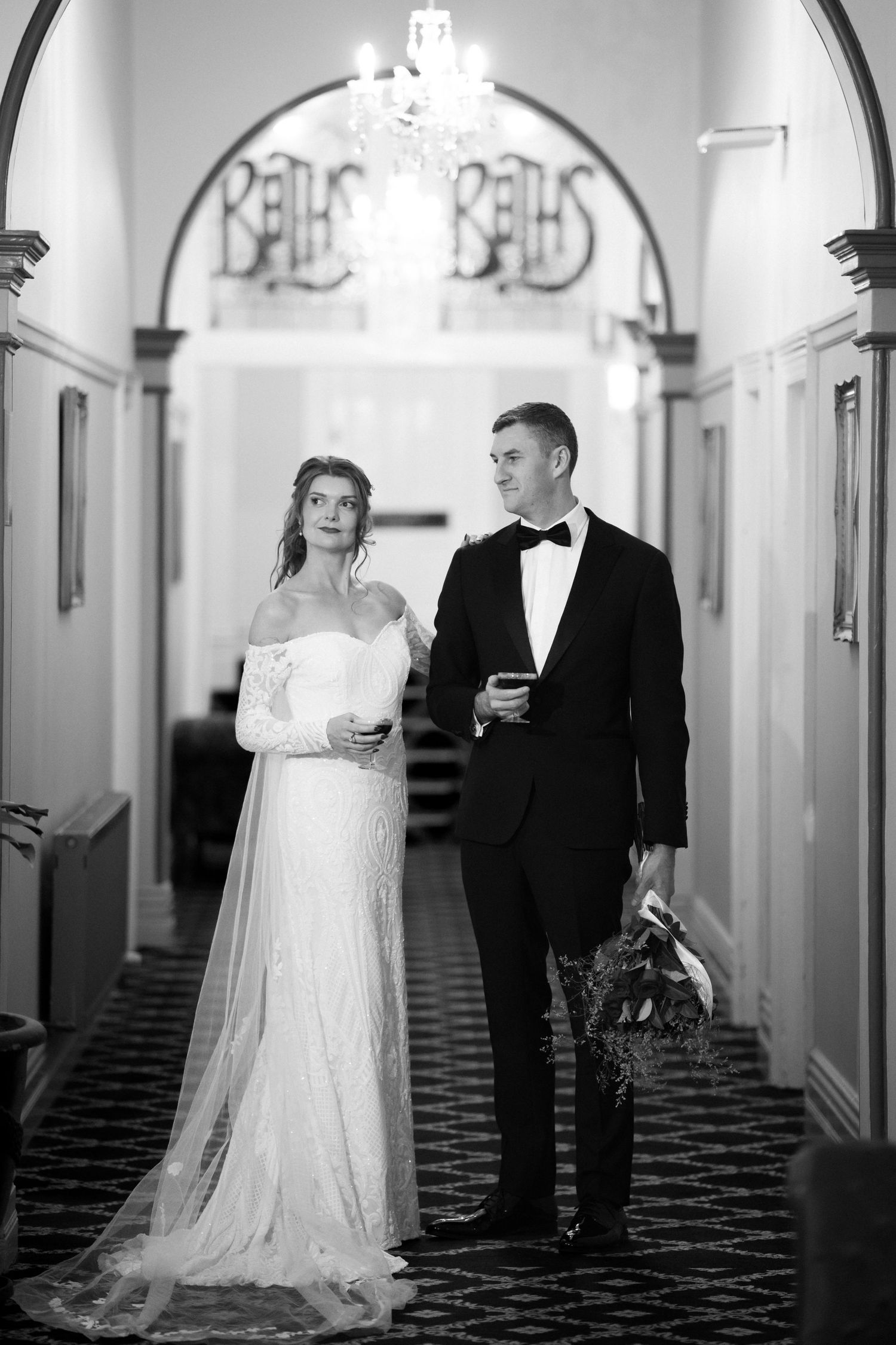 Black and white photo of bride in off-shoulder gown and groom in tuxedo standing in elegant hotel hallway.