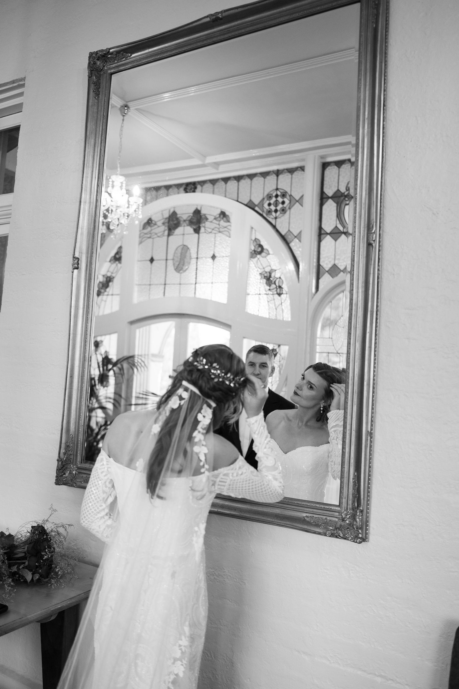 Bride in lace wedding dress adjusts hair accessory before ornate mirror with groom reflection in black and white.