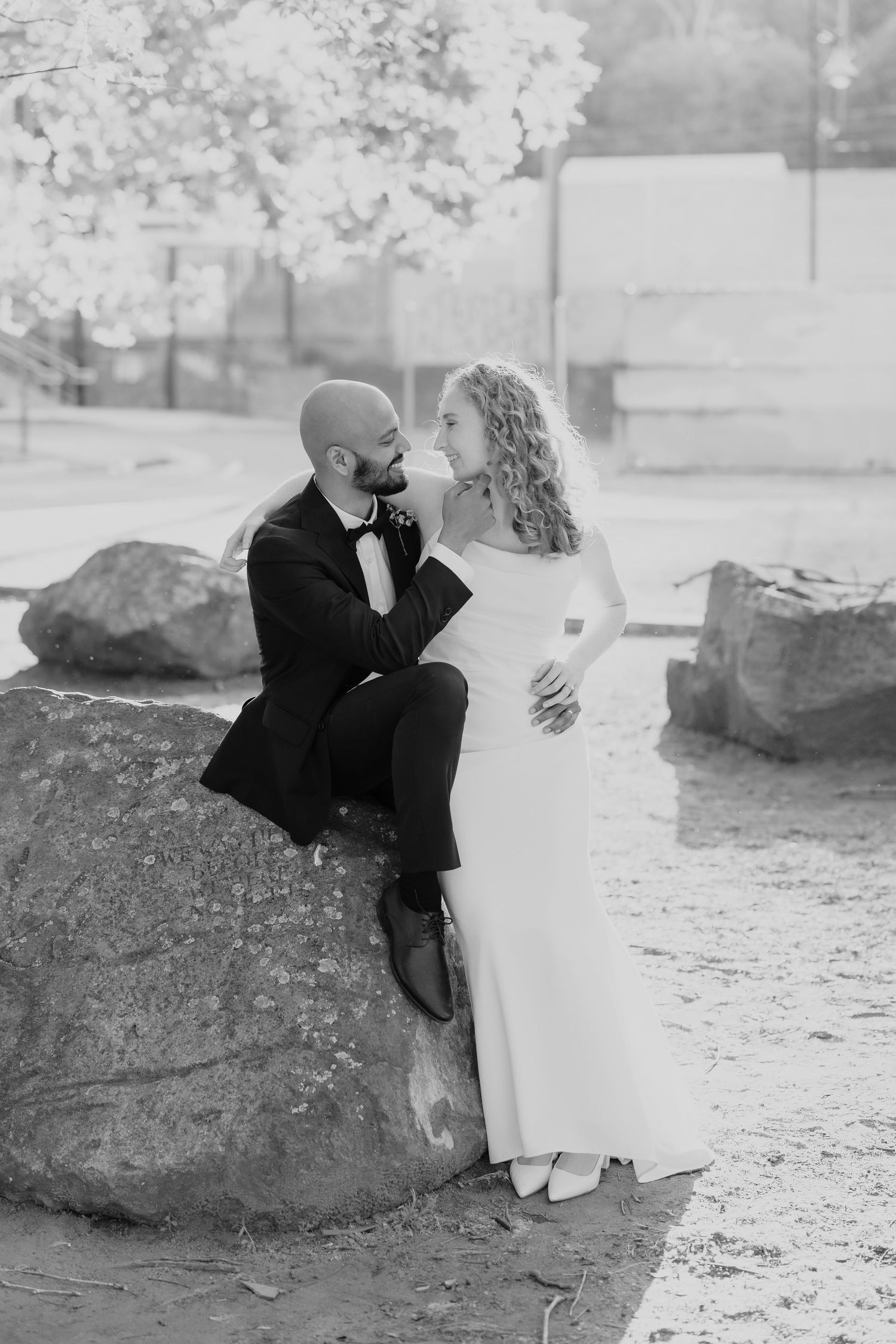 Black and white photo of a bride and groom sitting on a rock, gazing at each other lovingly on their wedding day.