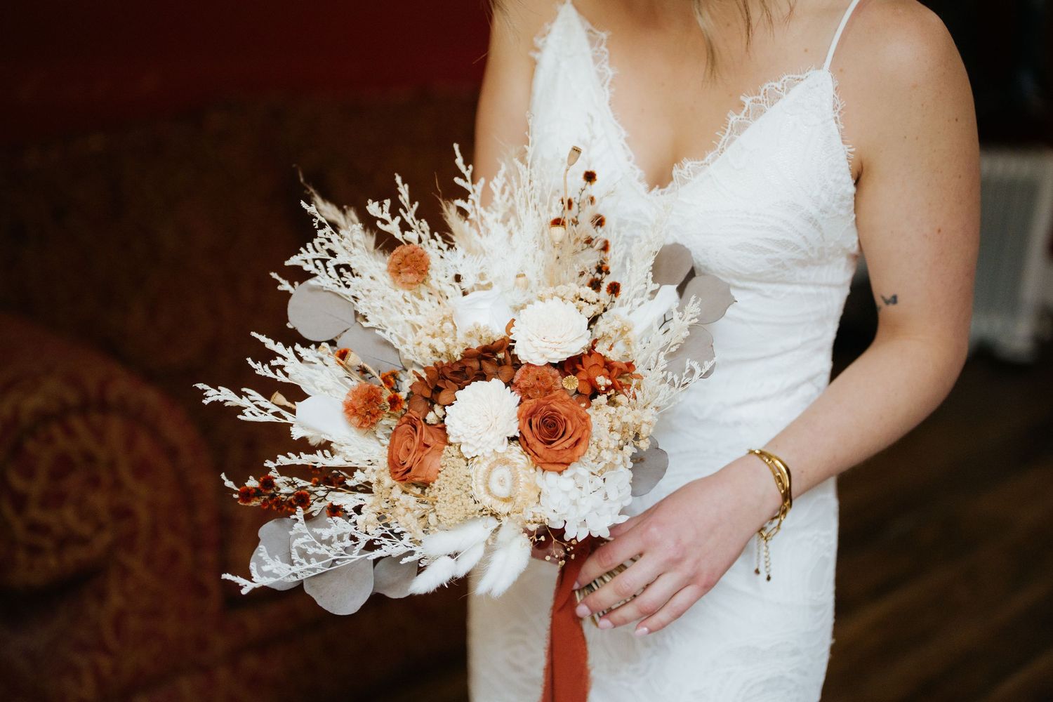 Bride in white lace gown holding a boho wedding bouquet with burnt orange roses, dried white florals, and pampas grass.