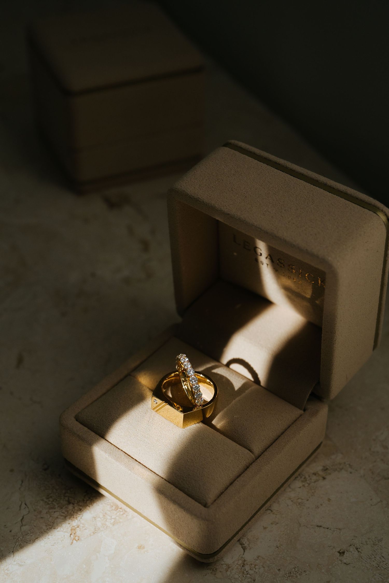 Gold wedding rings with diamonds displayed in an open beige velvet ring box, illuminated by warm sunlight.