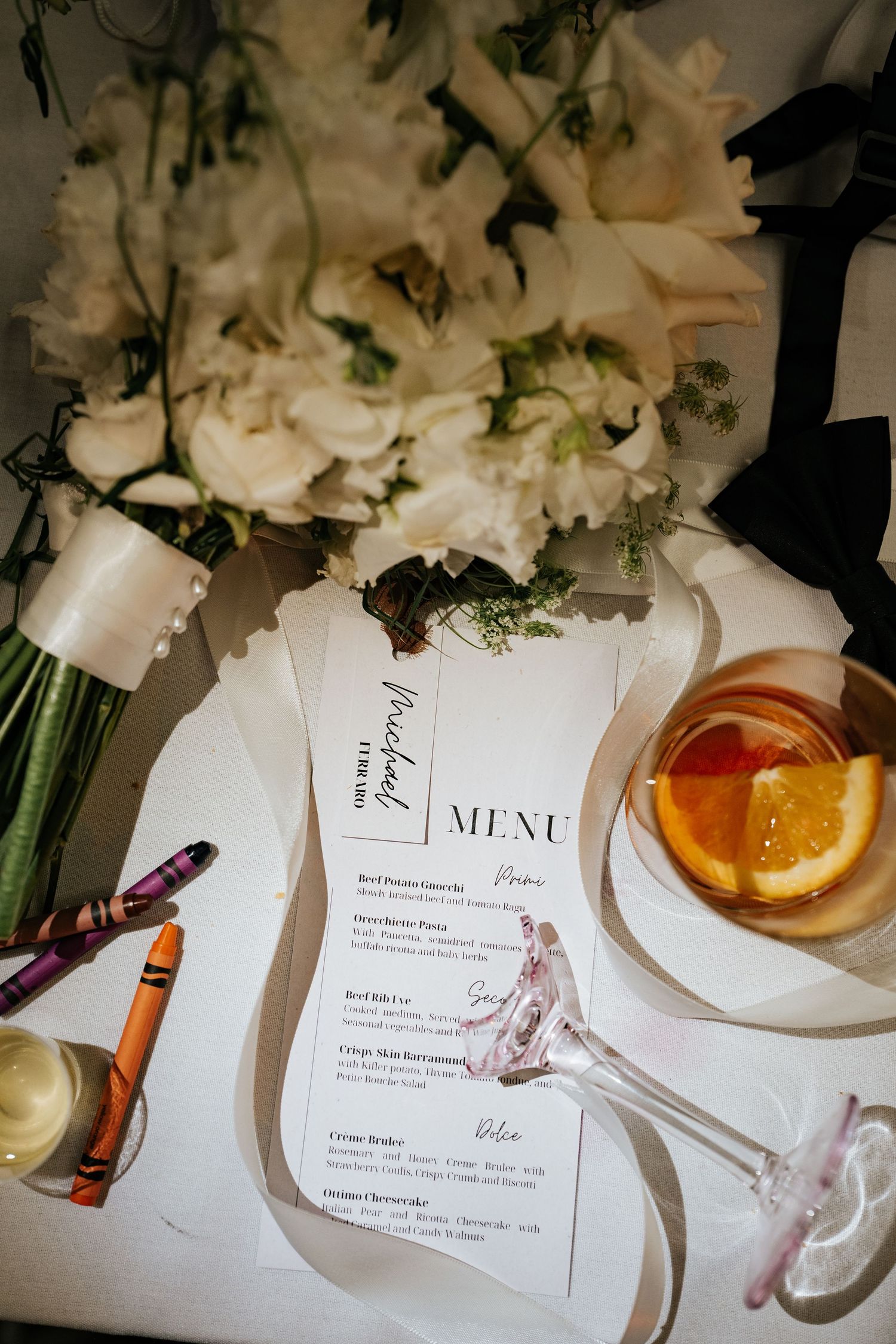 Elegant wedding menu card beside white floral bouquet, crayons, and cocktail with orange slice on table.