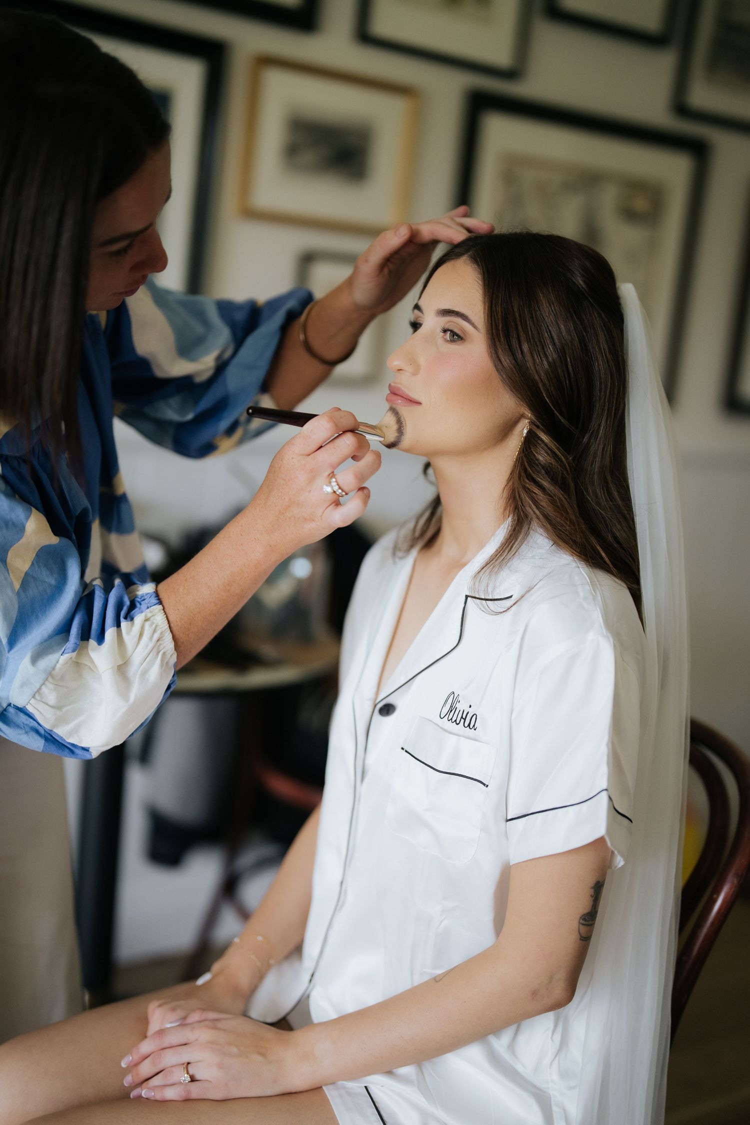 Makeup artist applying eyeshadow to brunette bride wearing white monogrammed pajamas and veil.