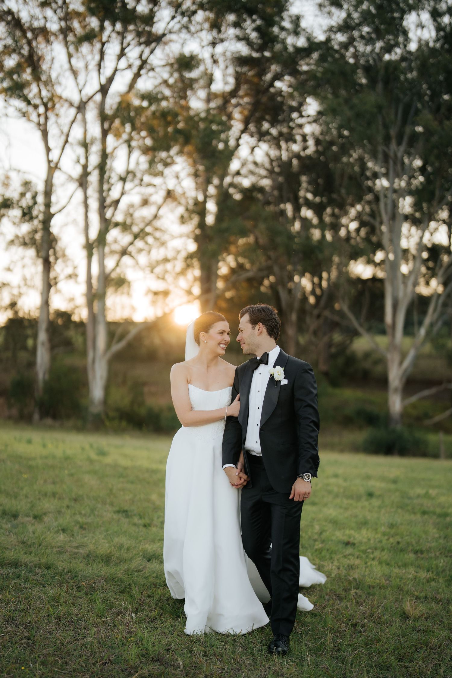 Bride and groom share a romantic sunset moment in a lush green field, with golden light glowing through eucalyptus trees.