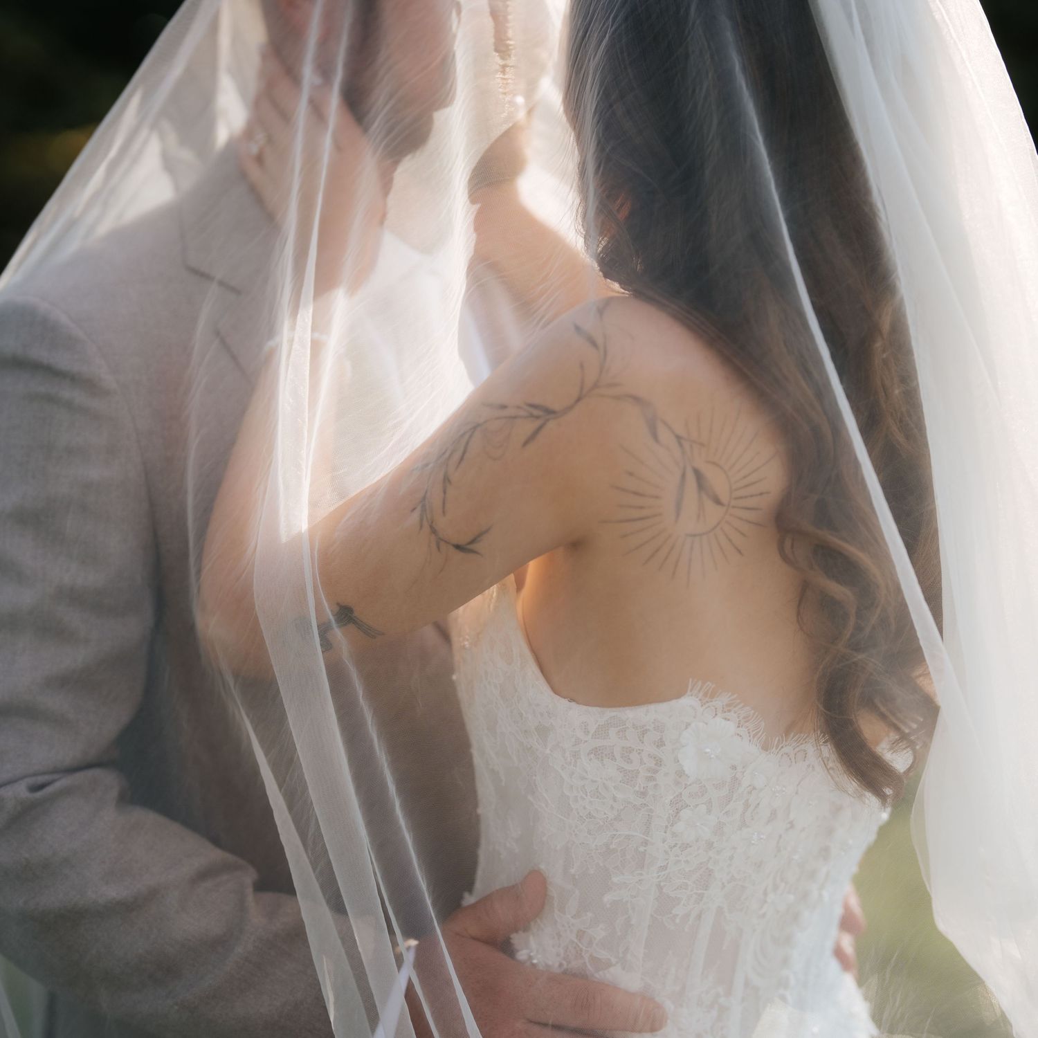 Bride and groom share an intimate moment under a veil, showcasing the bride's floral back tattoos and lace wedding dress.