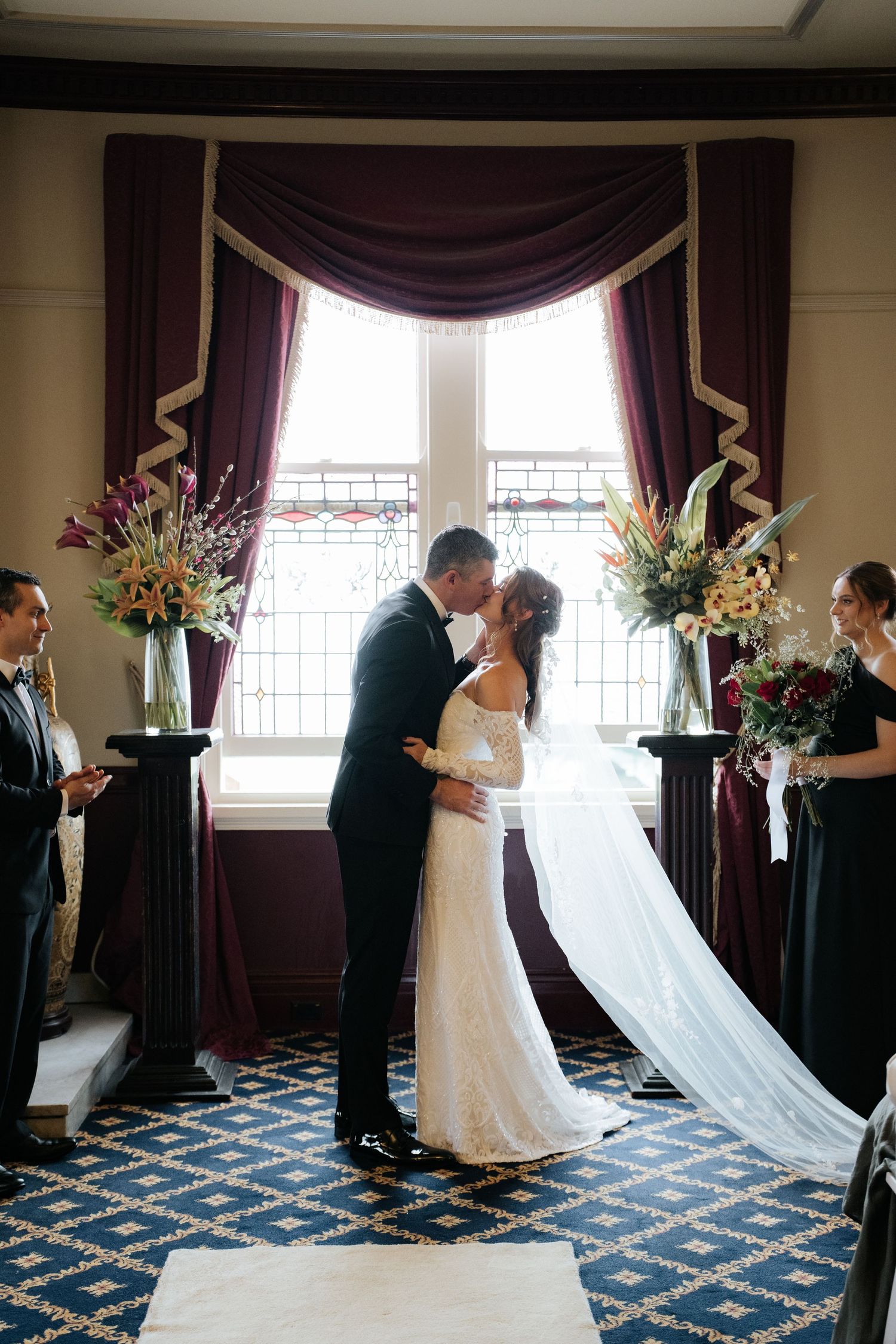 Bride and groom share first kiss at elegant wedding ceremony with burgundy drapes and floral arrangements.