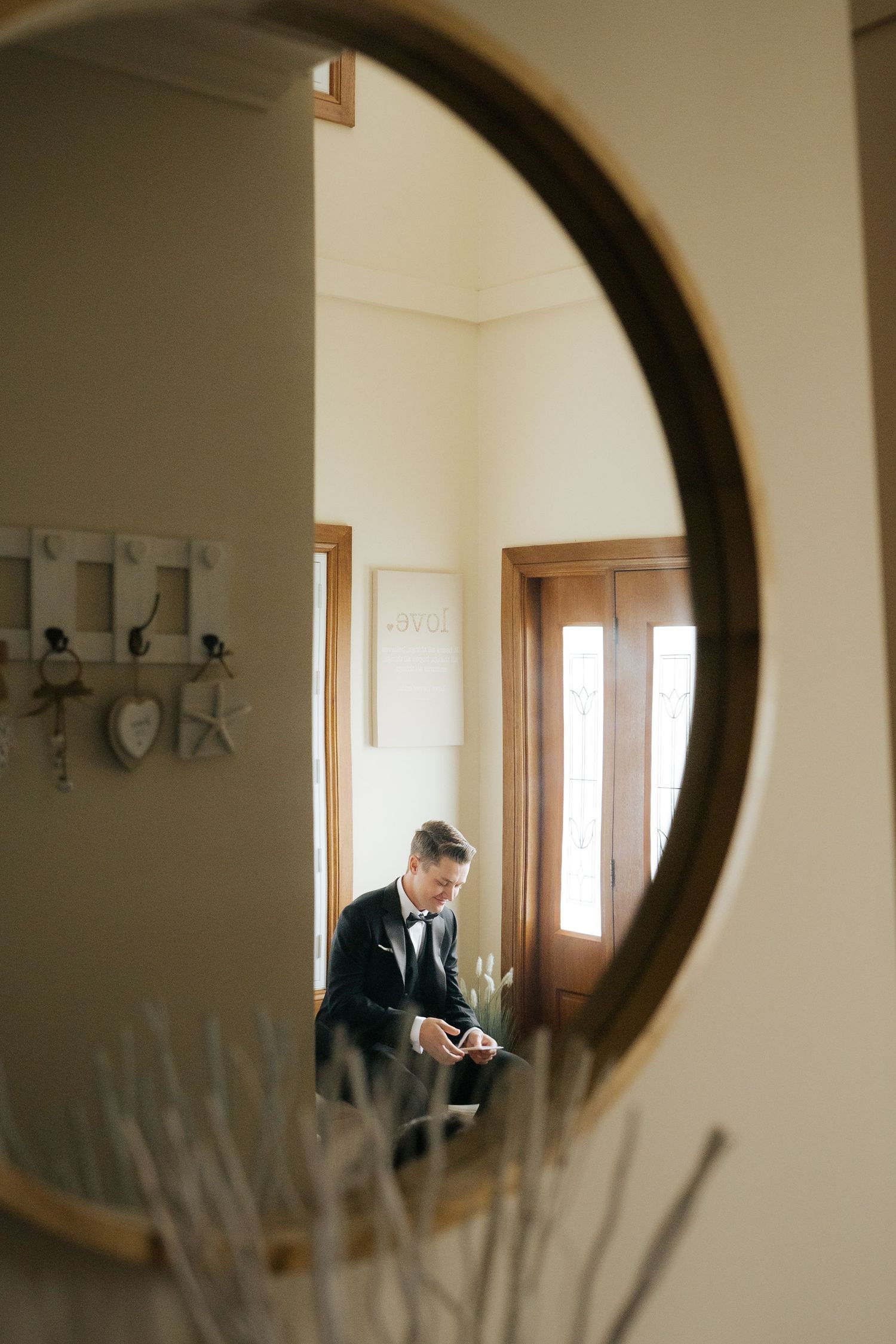 Groom in black tuxedo reflected in a round mirror while preparing for his wedding day.