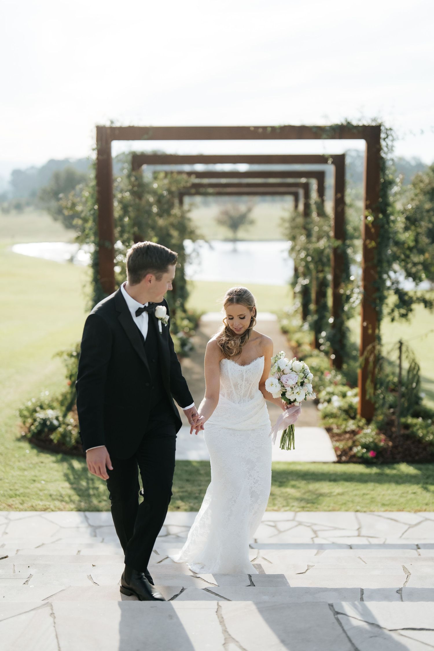 Bride and groom hold hands walking away from rustic wooden arch ceremony backdrop on lush green golf course.