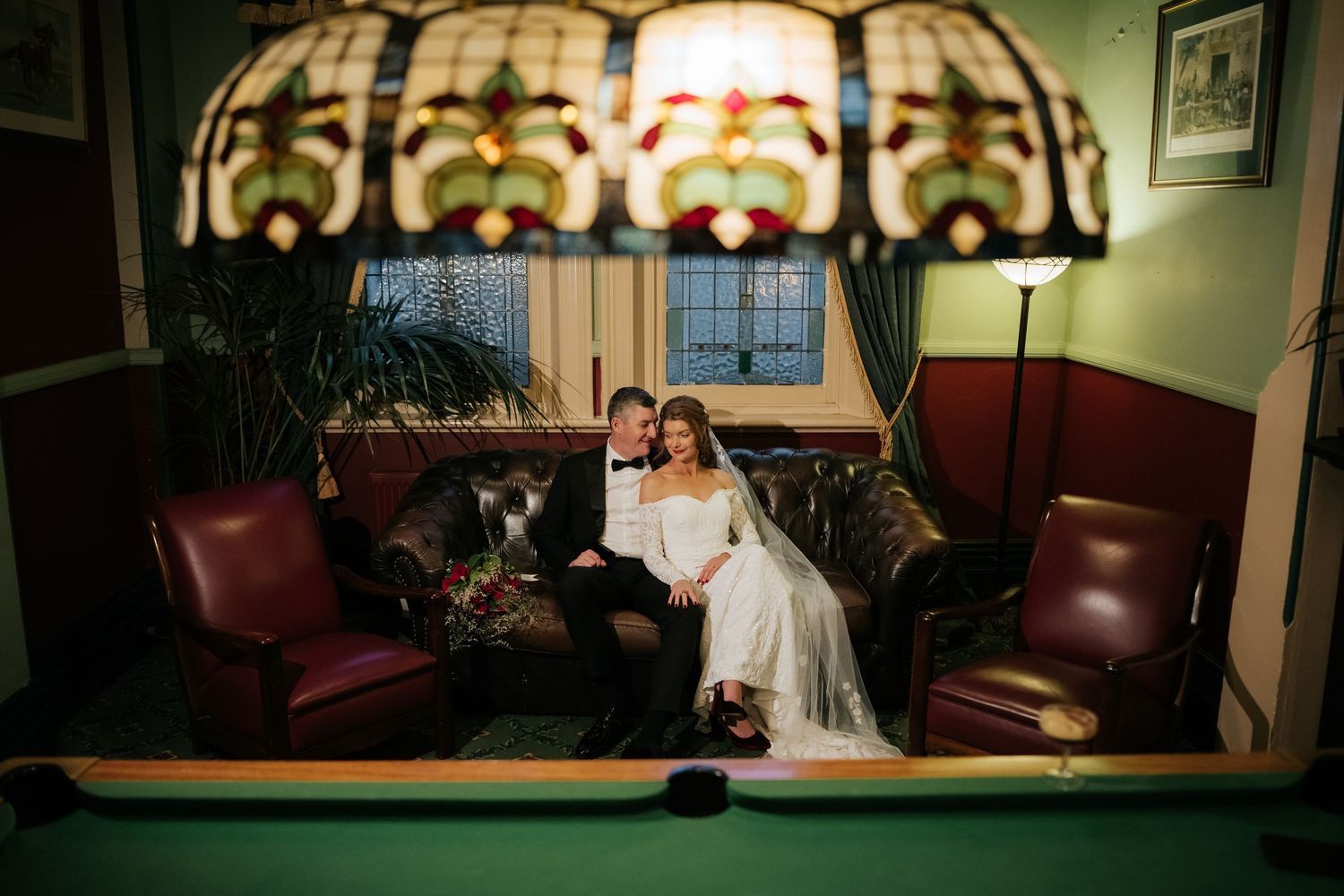 Bride and groom sitting together on a leather sofa under a stained glass lamp near a pool table.
