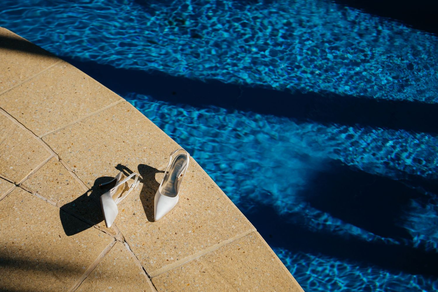 White bridal heels and accessories placed on a sunny poolside terrace beside a shimmering blue swimming pool.
