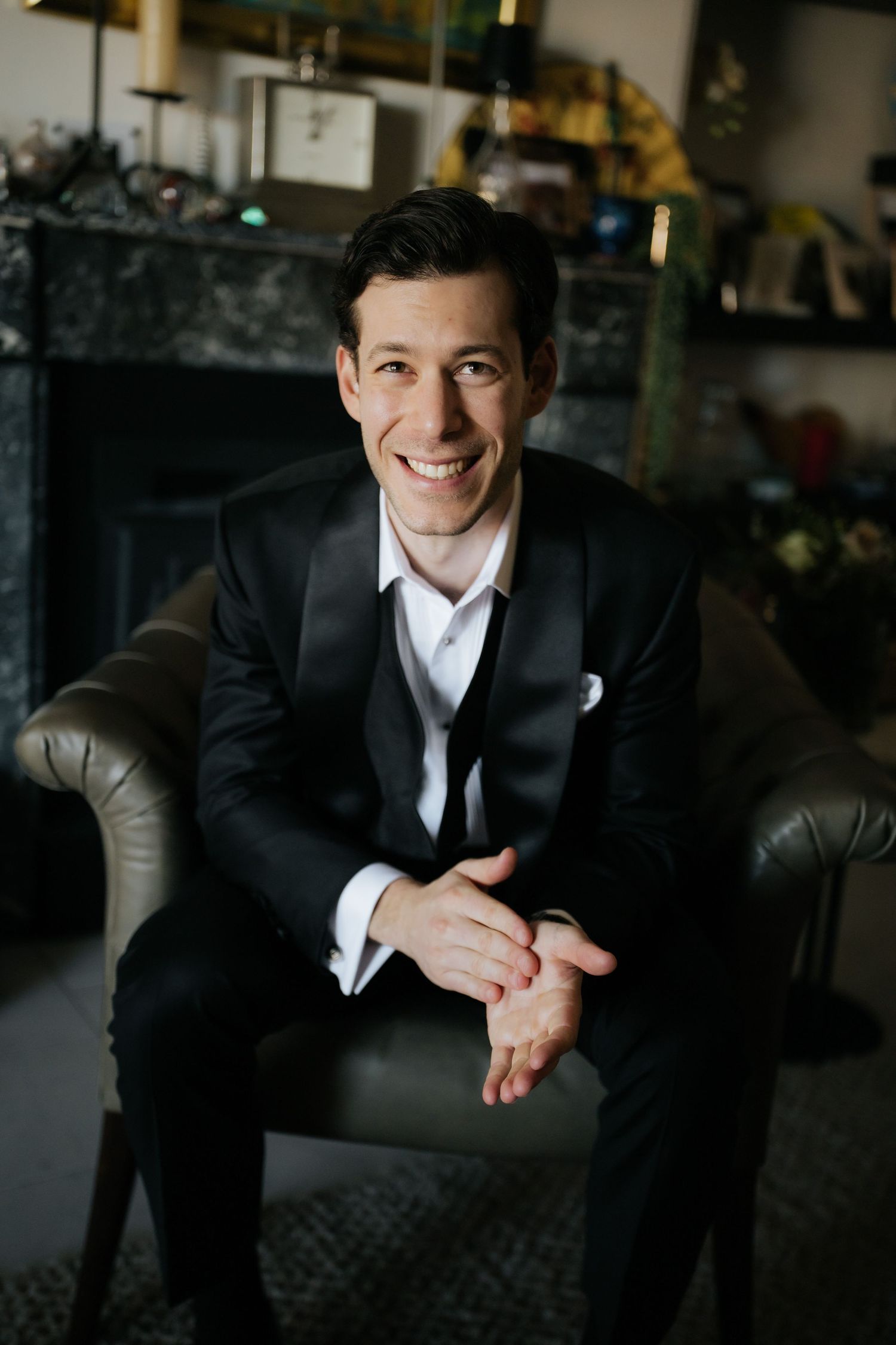 Smiling groom in black tuxedo sitting in leather chair with clasped hands at elegant indoor wedding venue.