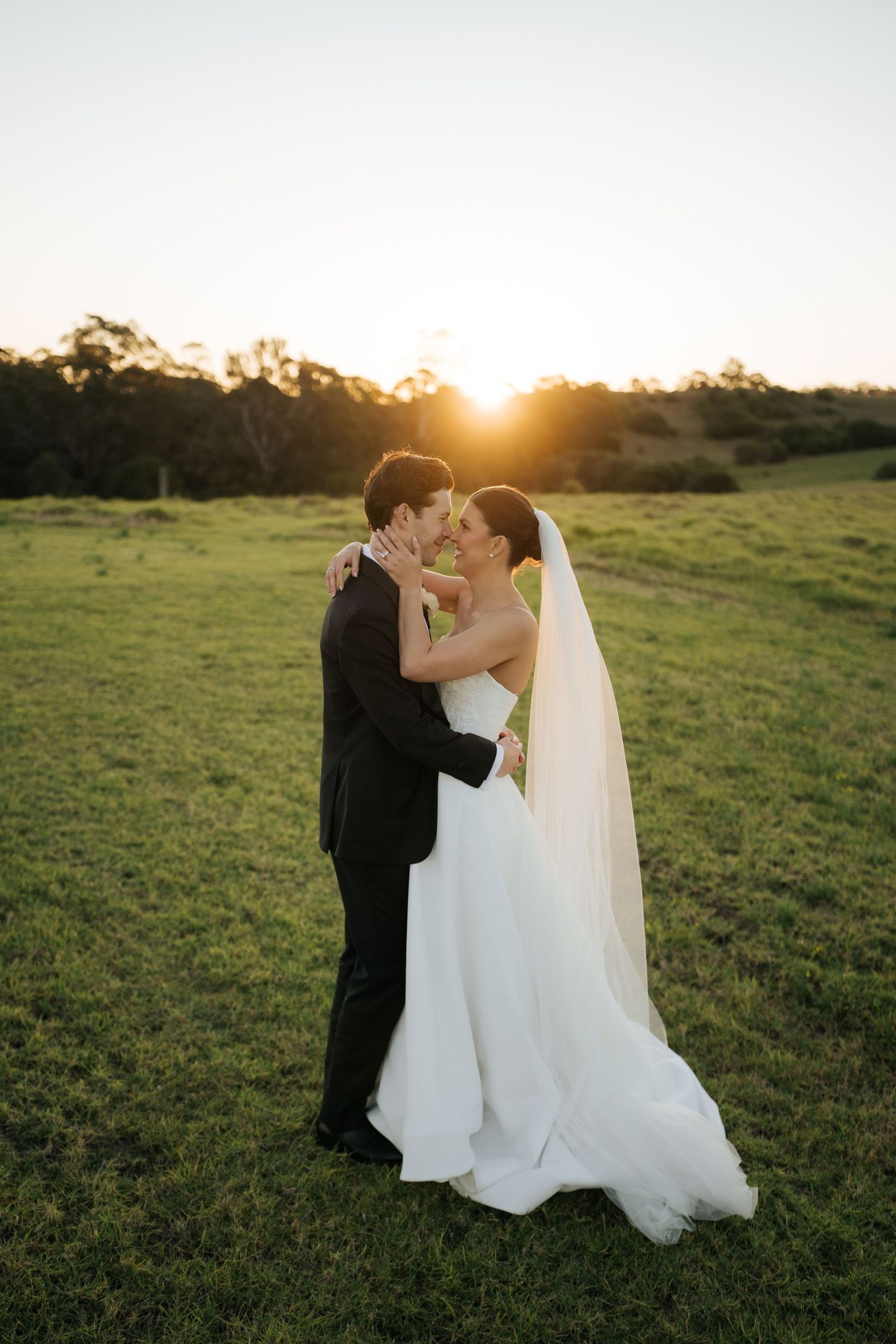 Bride and groom sharing romantic embrace in open field during stunning golden sunset.
