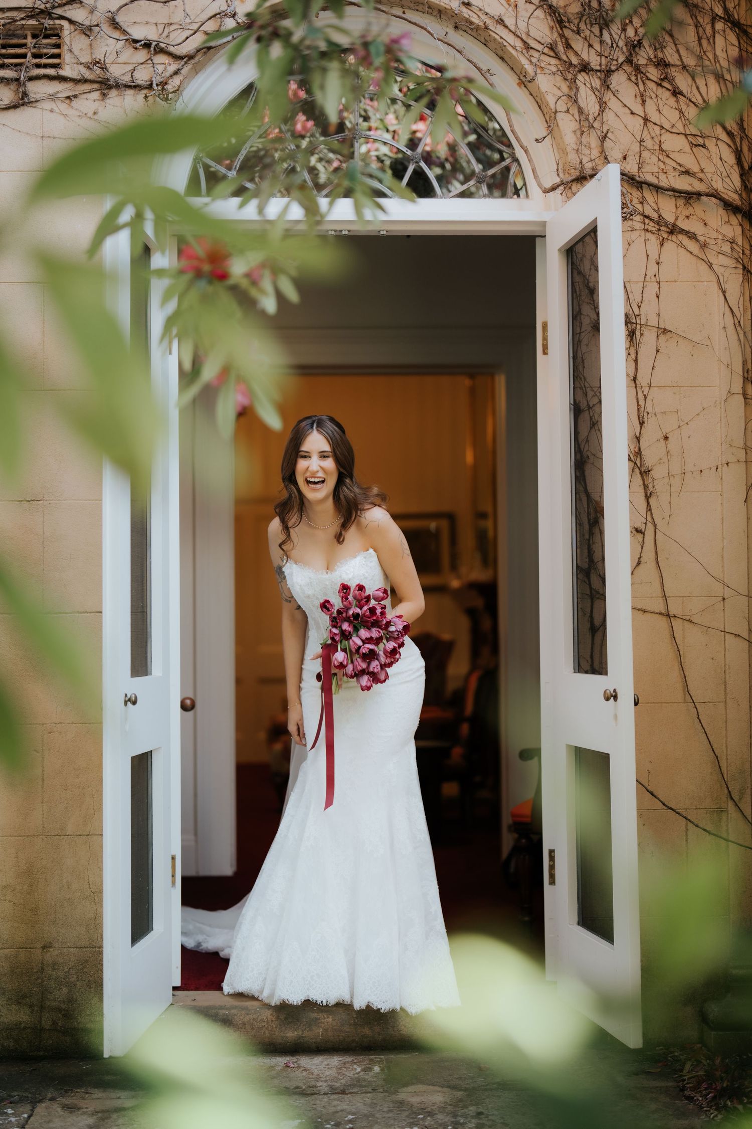 Bride in white mermaid gown holding red bouquet, standing in open French doors of elegant stone venue.