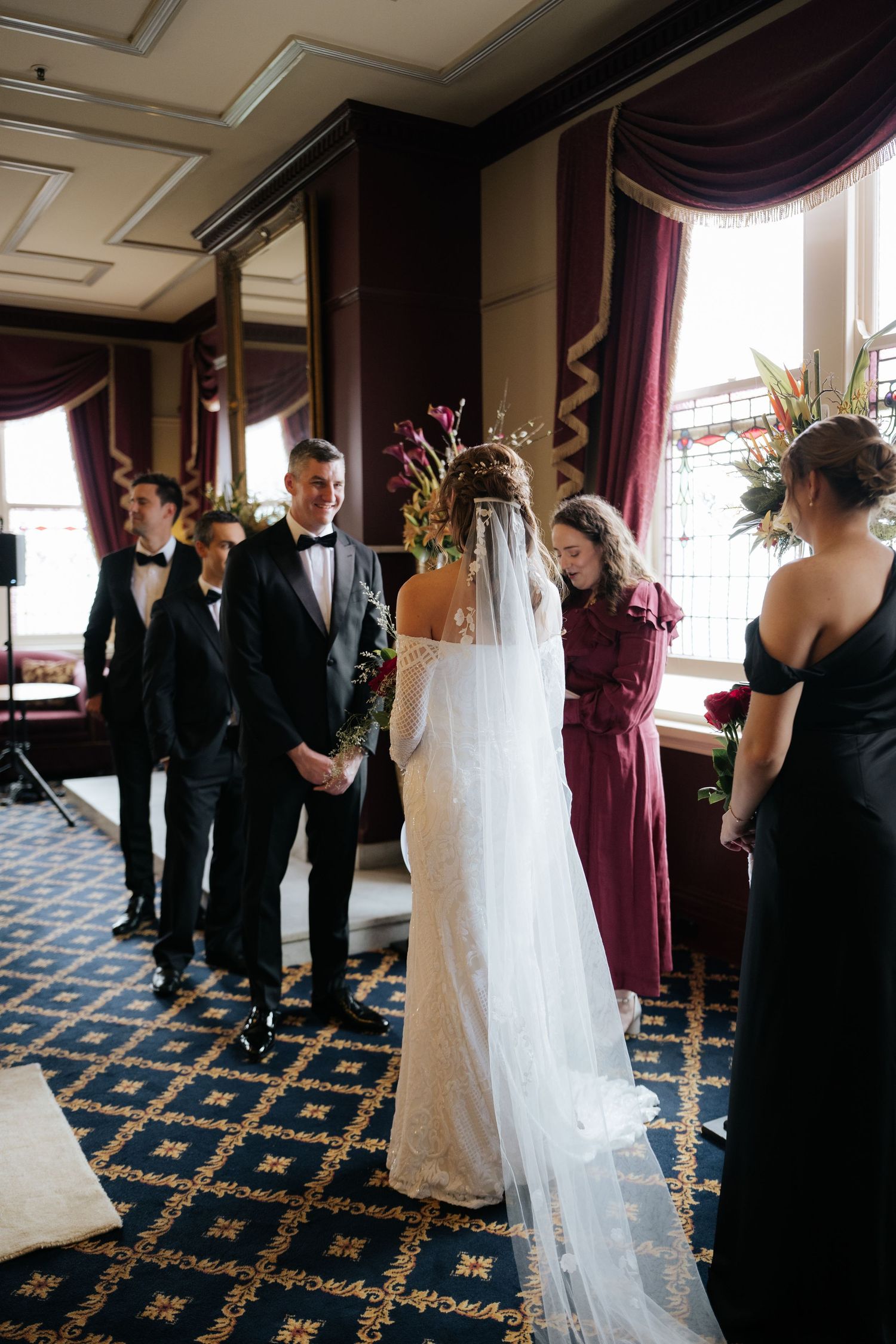 Bride and groom exchange vows at elegant indoor wedding ceremony with burgundy drapes and floral arrangements.