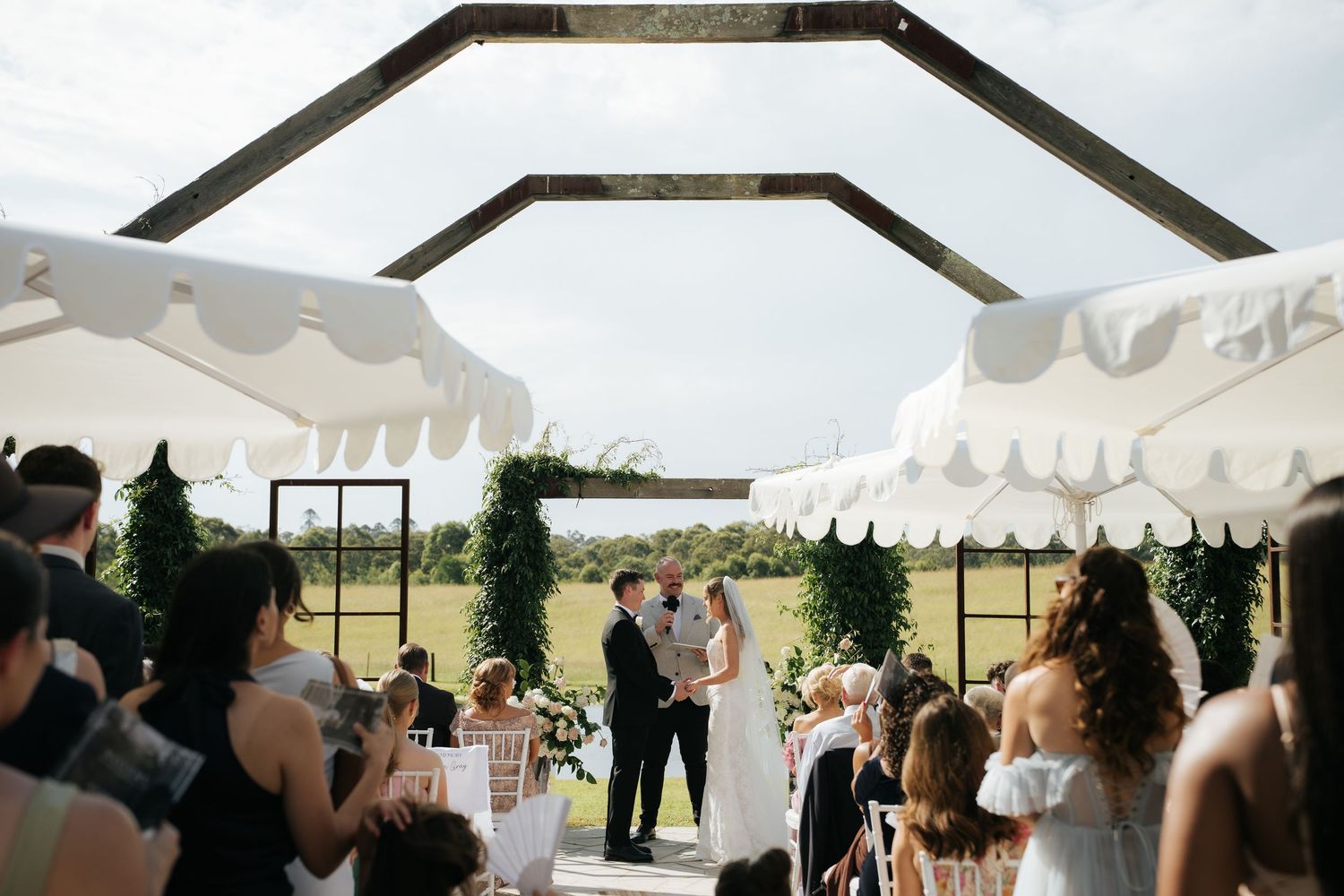 Outdoor wedding ceremony under geometric pergola with white striped umbrellas, couple kissing at altar surrounded by greenery.