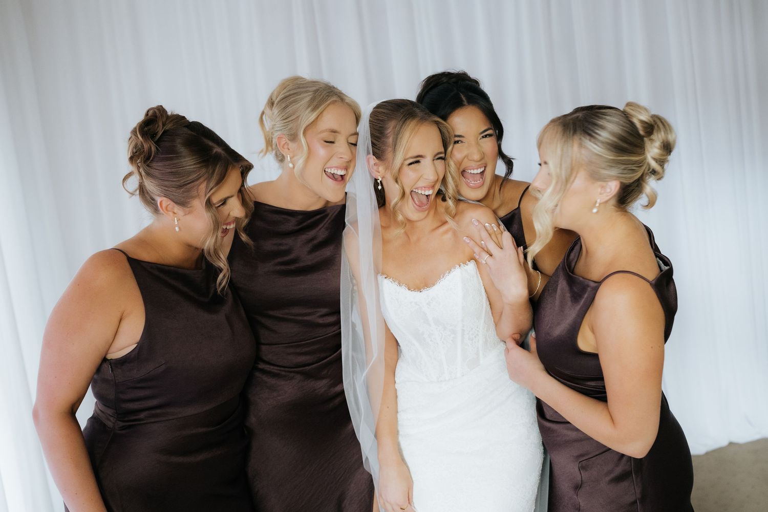 Bride in white strapless gown laughing with four bridesmaids in dark brown satin dresses during wedding preparations.