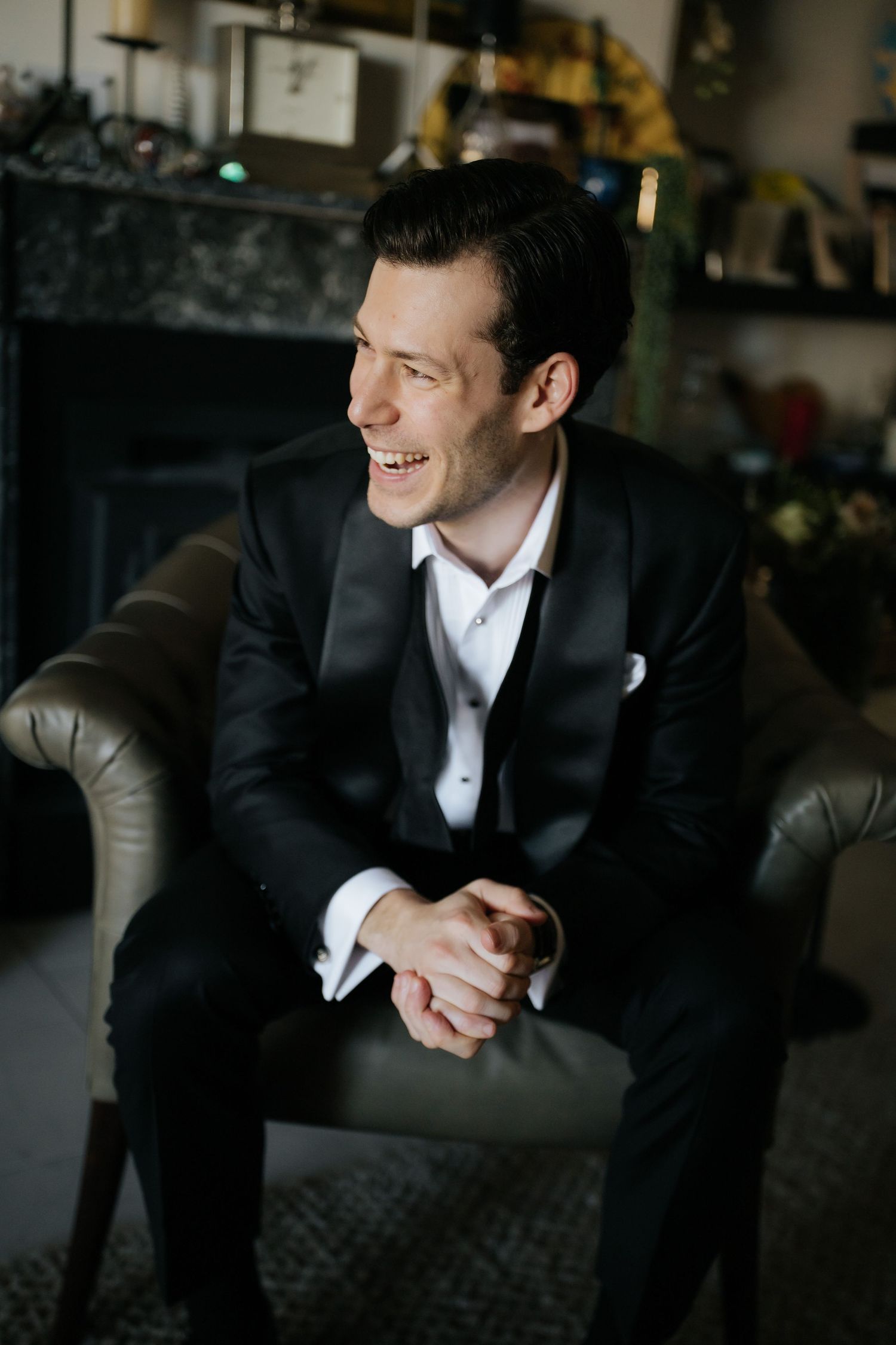 Smiling groom in black tuxedo sitting in leather chair at wedding venue.