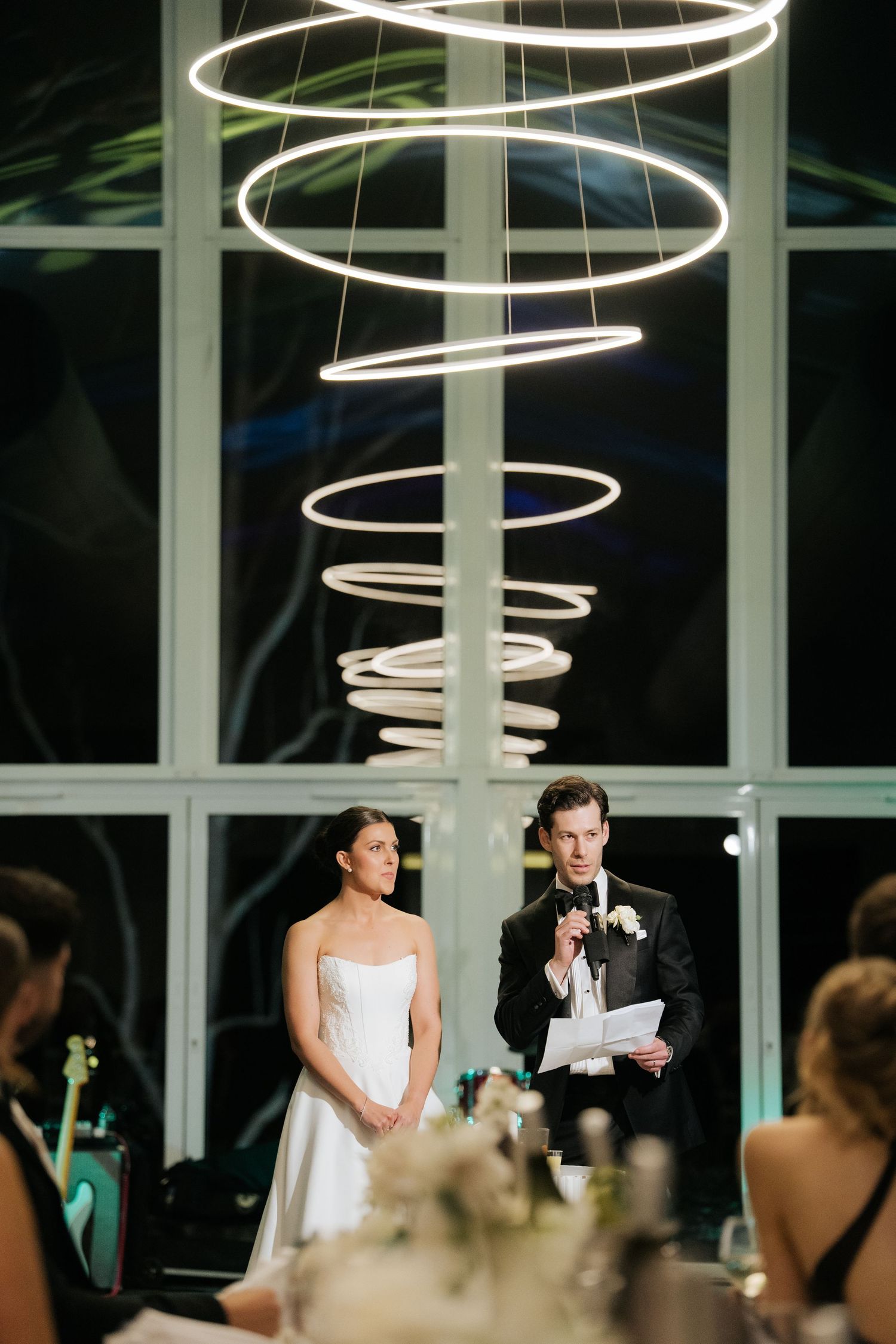 Bride and groom stand beneath modern ring chandelier during wedding reception speech in elegant venue.