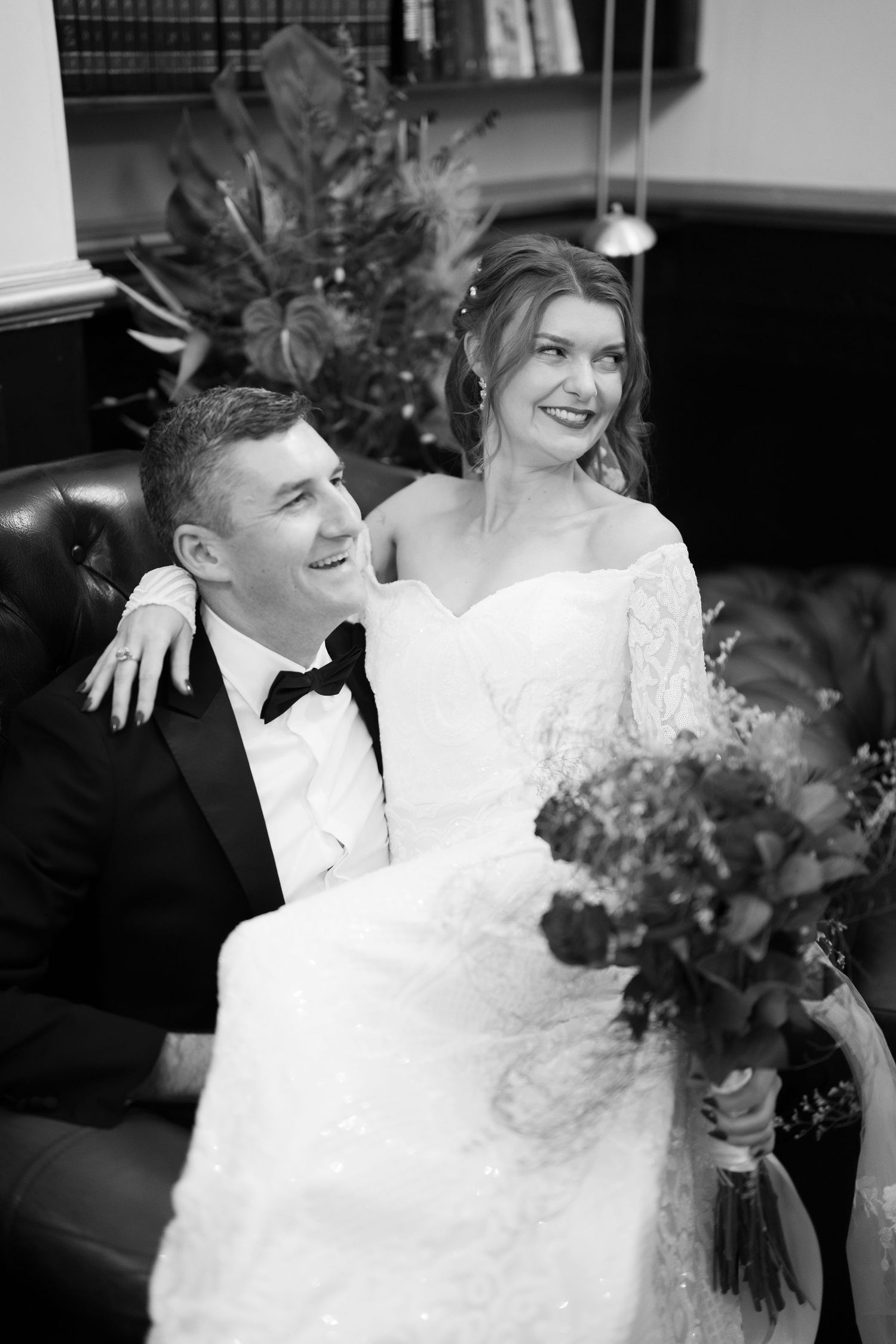 Black and white photo of a smiling bride and groom sitting together, bride holding a bouquet, groom in a tuxedo.