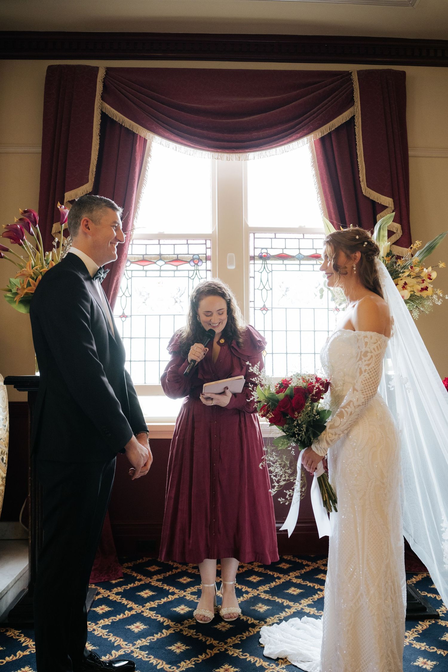 Bride and groom exchange vows with officiant in burgundy dress, elegant room with draped windows and floral arrangements.