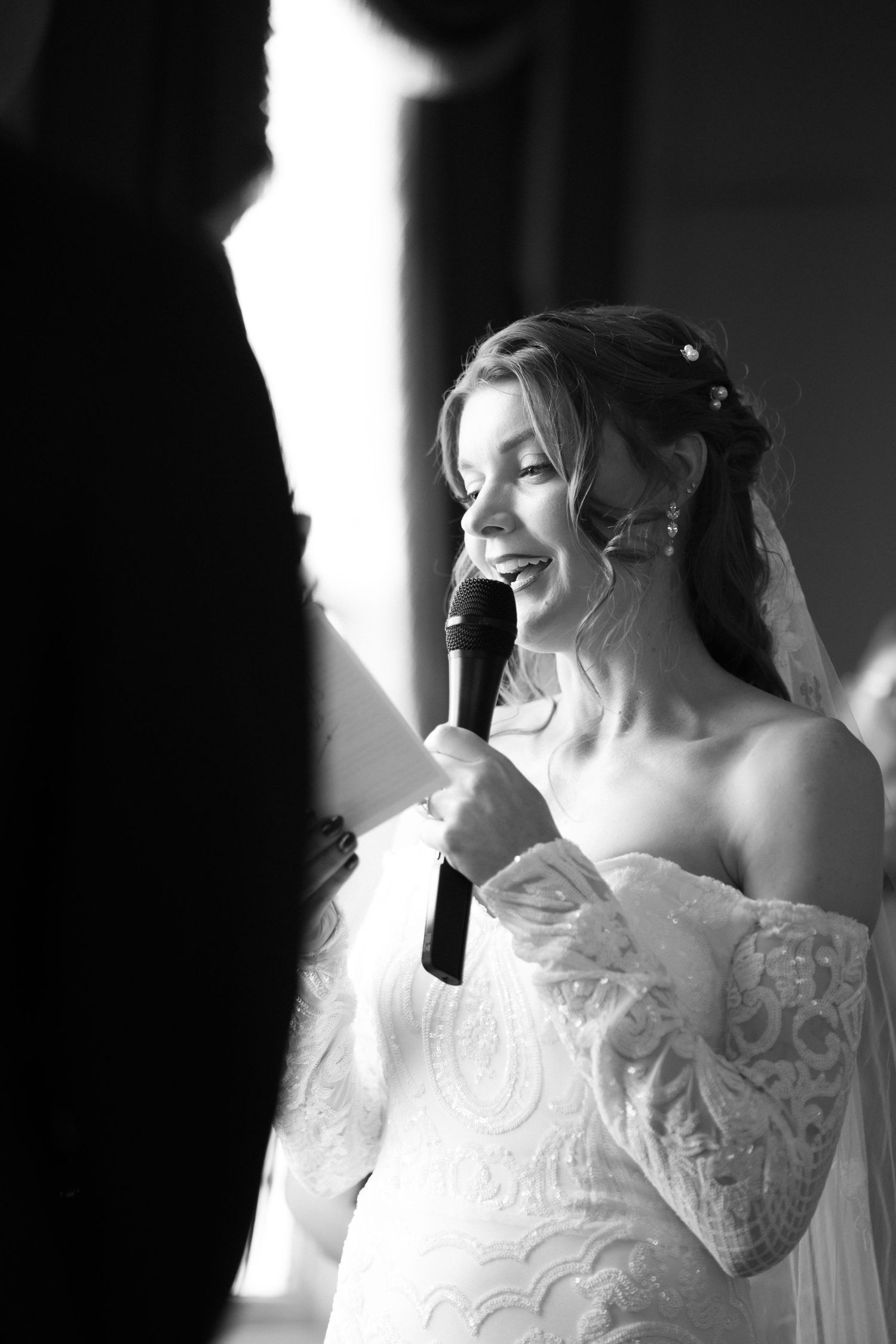 Bride in lace off-shoulder gown speaks into microphone, smiling during wedding ceremony, black and white photo.