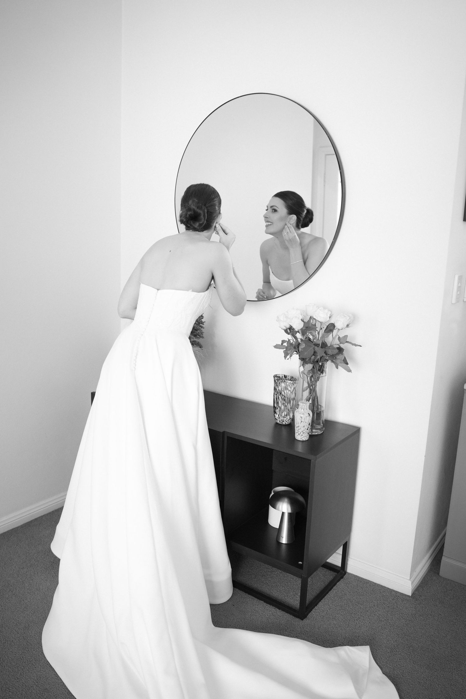 Bride in strapless white gown checks makeup in round mirror beside floral arrangement in black and white photo.