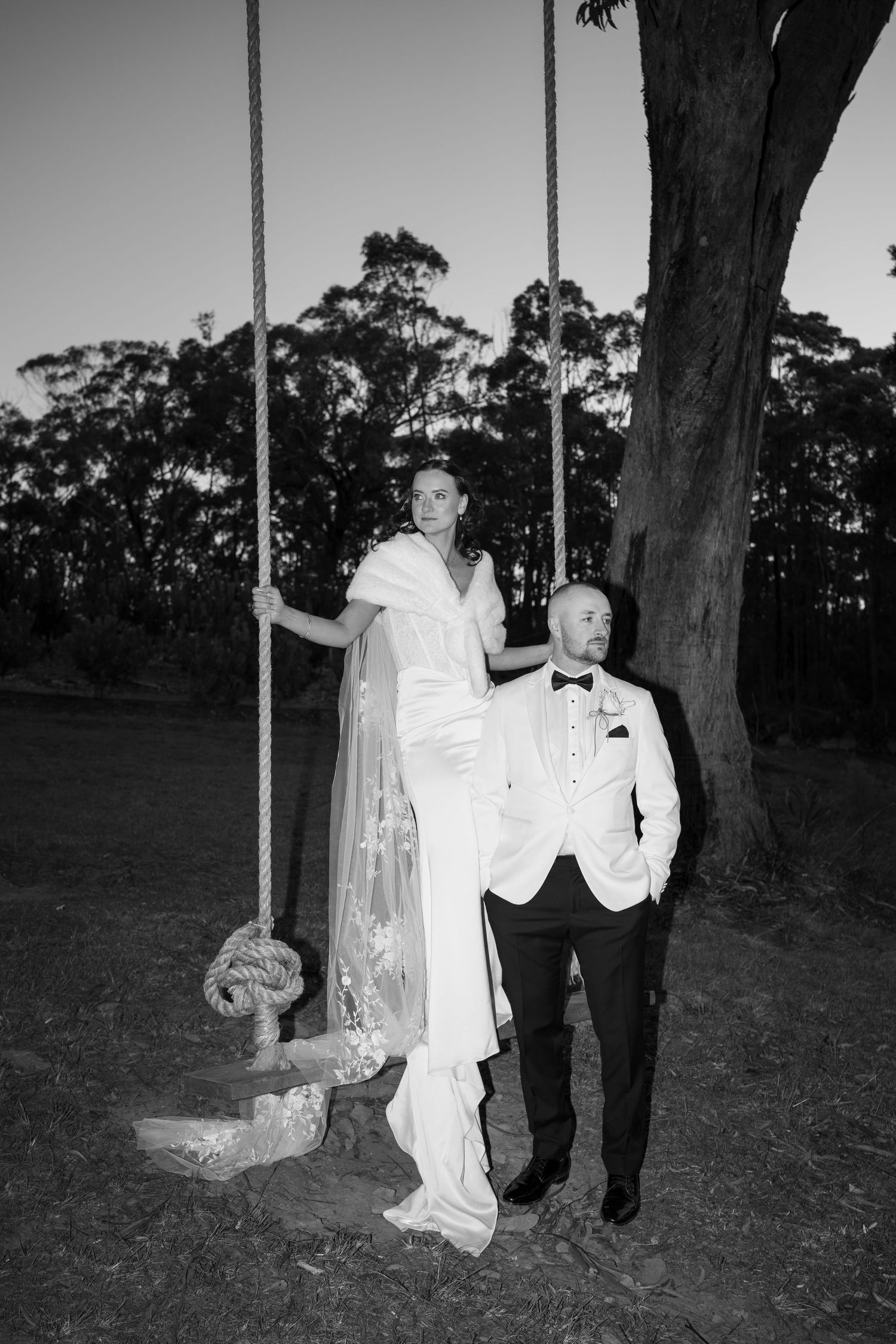 Bride and groom pose by a rope swing under a large tree at dusk in black and white photo.