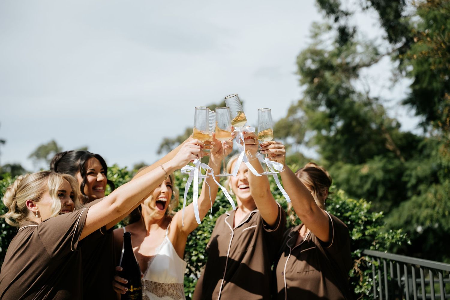 Bridesmaids in brown robes toasting champagne glasses outdoors at a wedding celebration.
