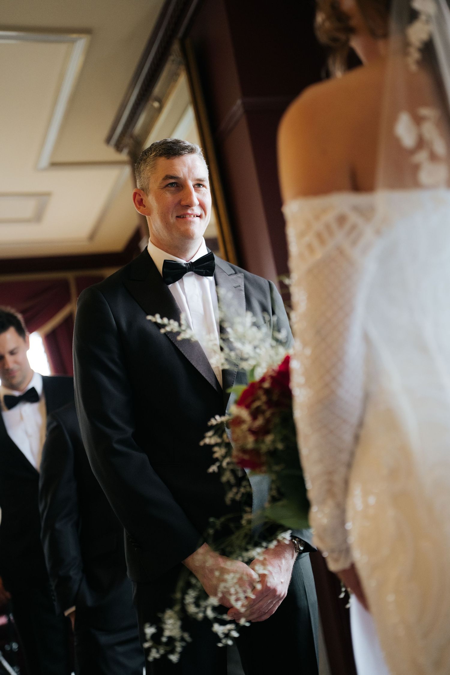 Groom in black tuxedo smiling at bride during wedding ceremony, with floral bouquet and elegant venue backdrop.