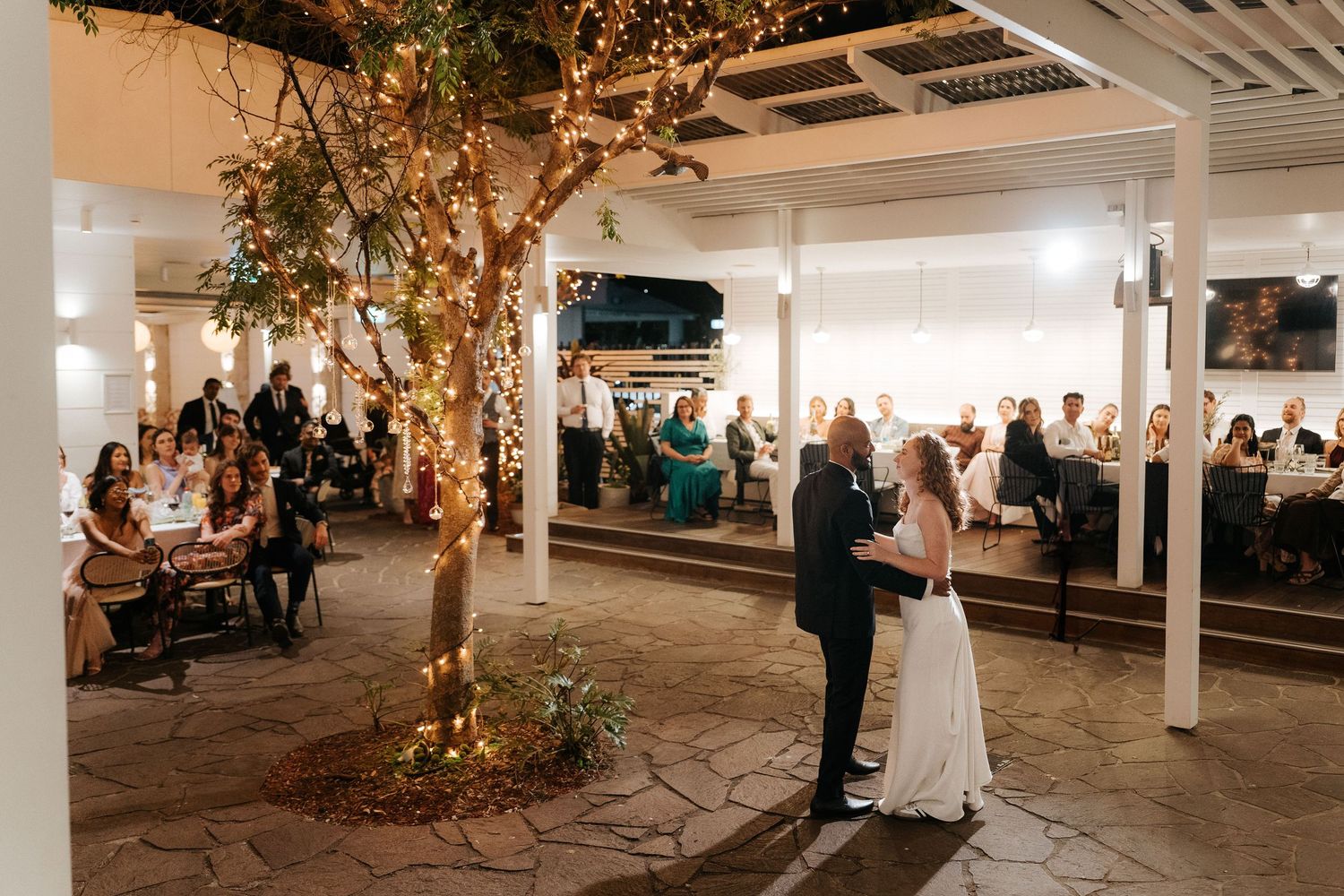 Couple shares first dance surrounded by guests at elegant indoor wedding venue with fairy lights on trees.