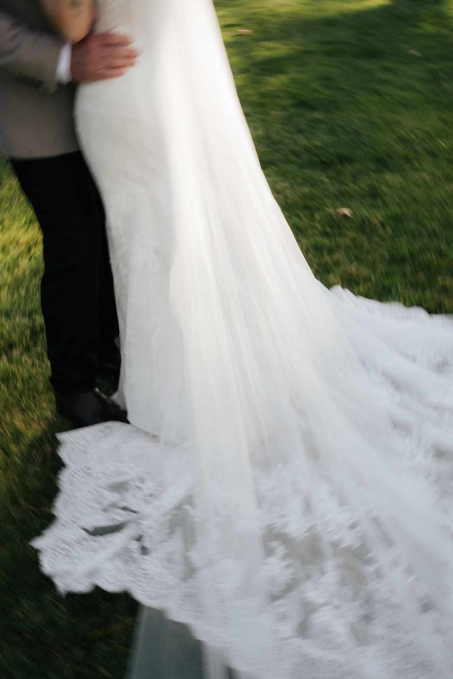 Bride in elegant lace wedding gown with long train standing with groom on green grass outdoor ceremony.