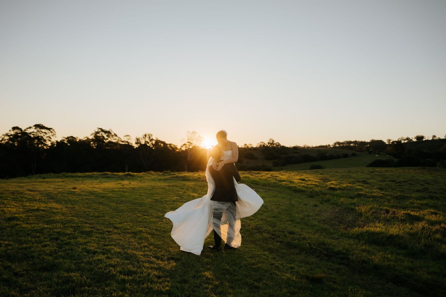Couple dancing at sunset in a green field, bride's white dress flowing in the wind during golden hour outdoor wedding.