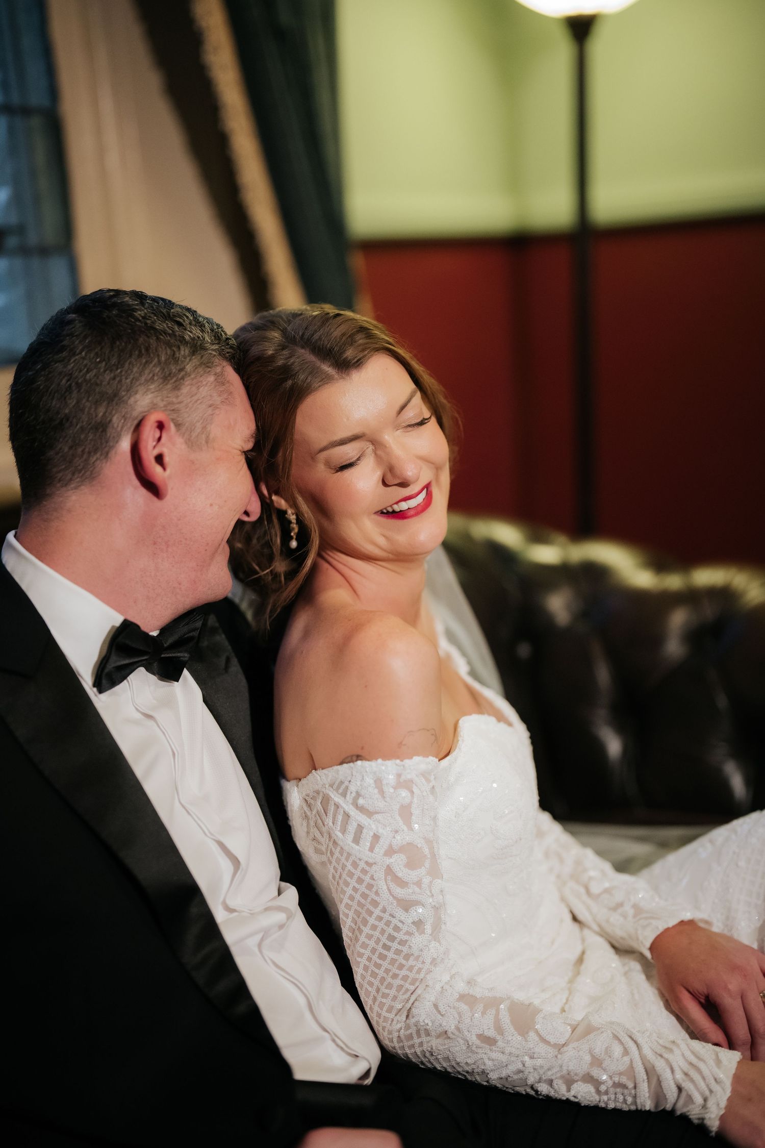 A bride in a lace off-shoulder gown laughs joyfully beside her groom in a black tuxedo on a leather sofa.