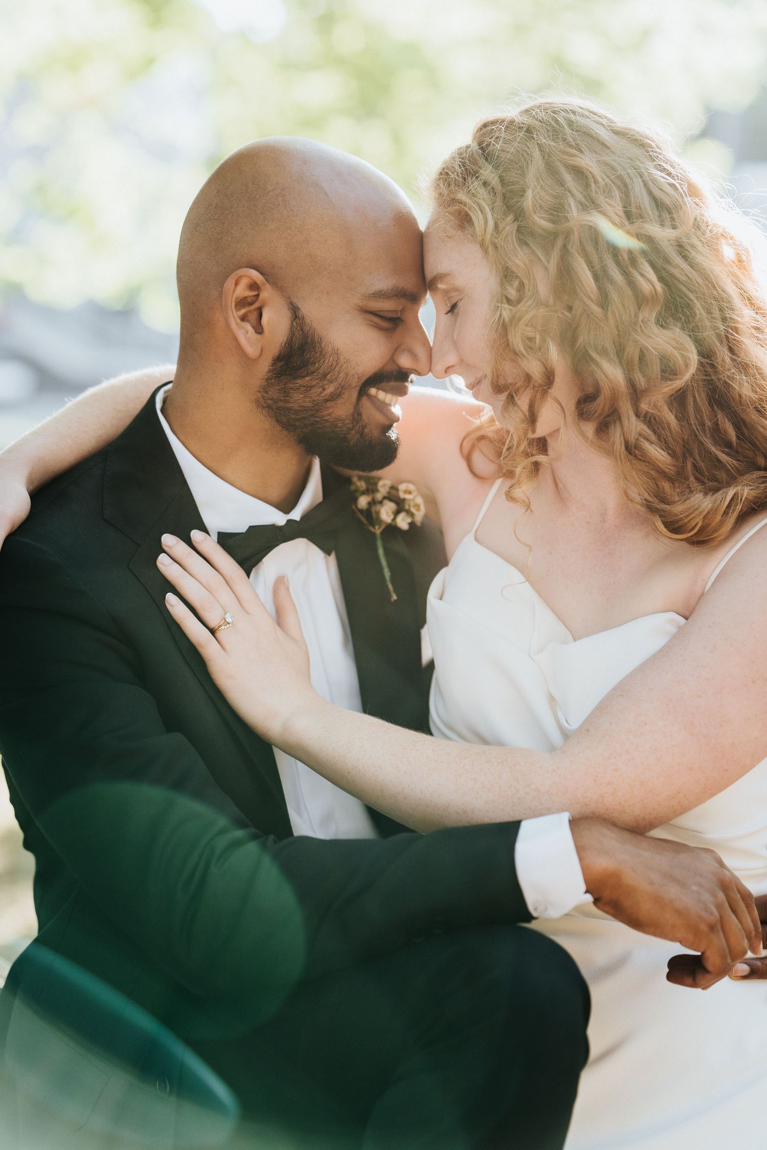 Interracial couple in wedding attire sharing a romantic forehead touch, smiling joyfully outdoors.