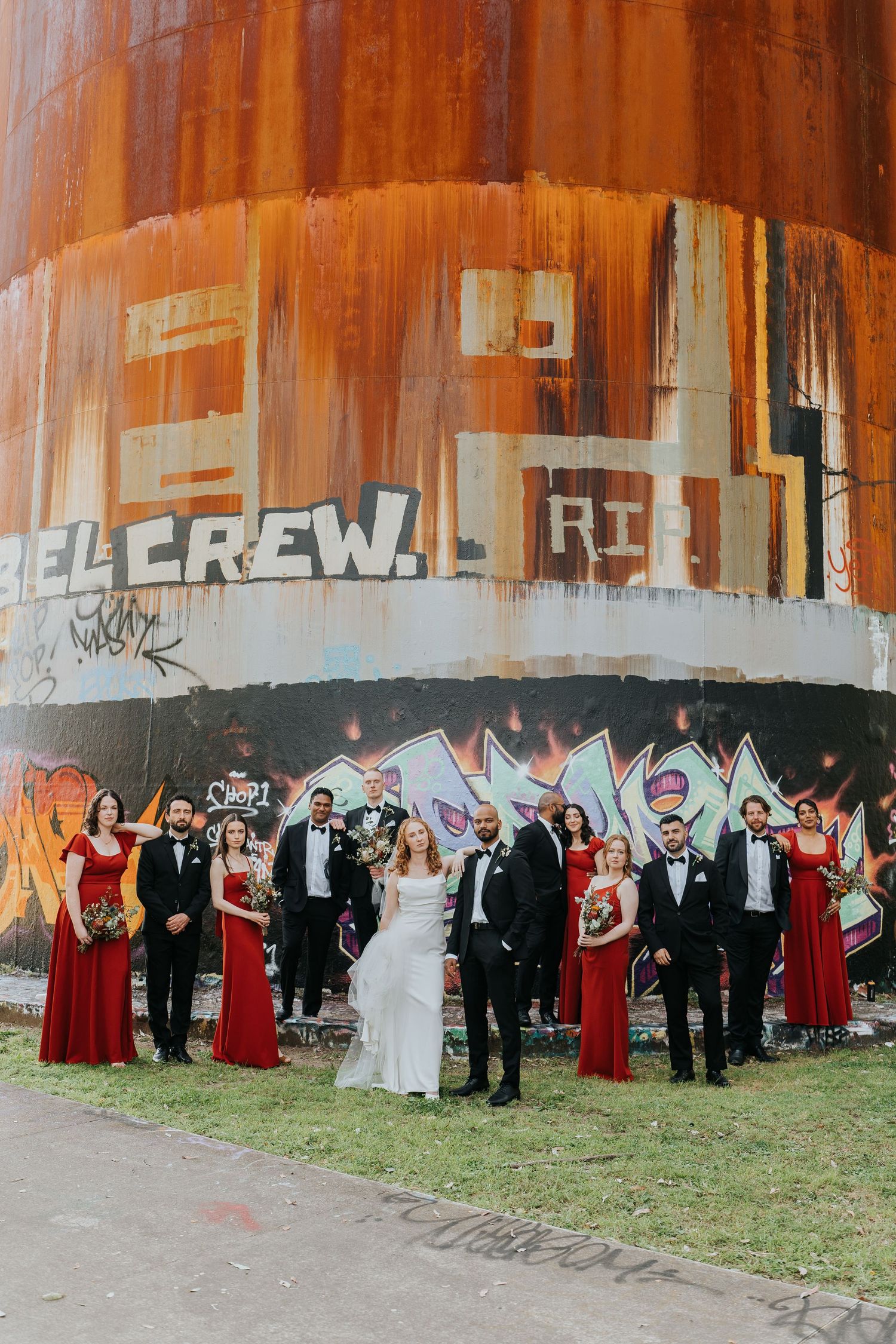 Wedding party in red bridesmaid dresses and tuxedos posing before a rusty graffiti-covered industrial silo.