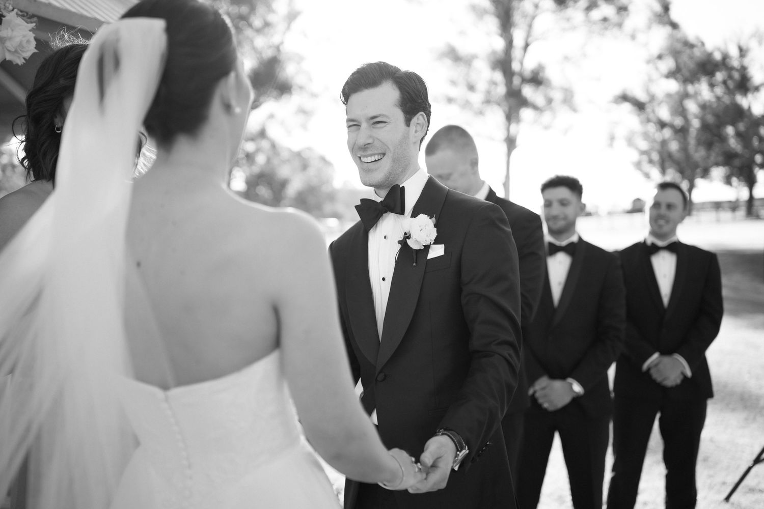 Black and white photo of a groom smiling at his bride during wedding ceremony with groomsmen standing behind.