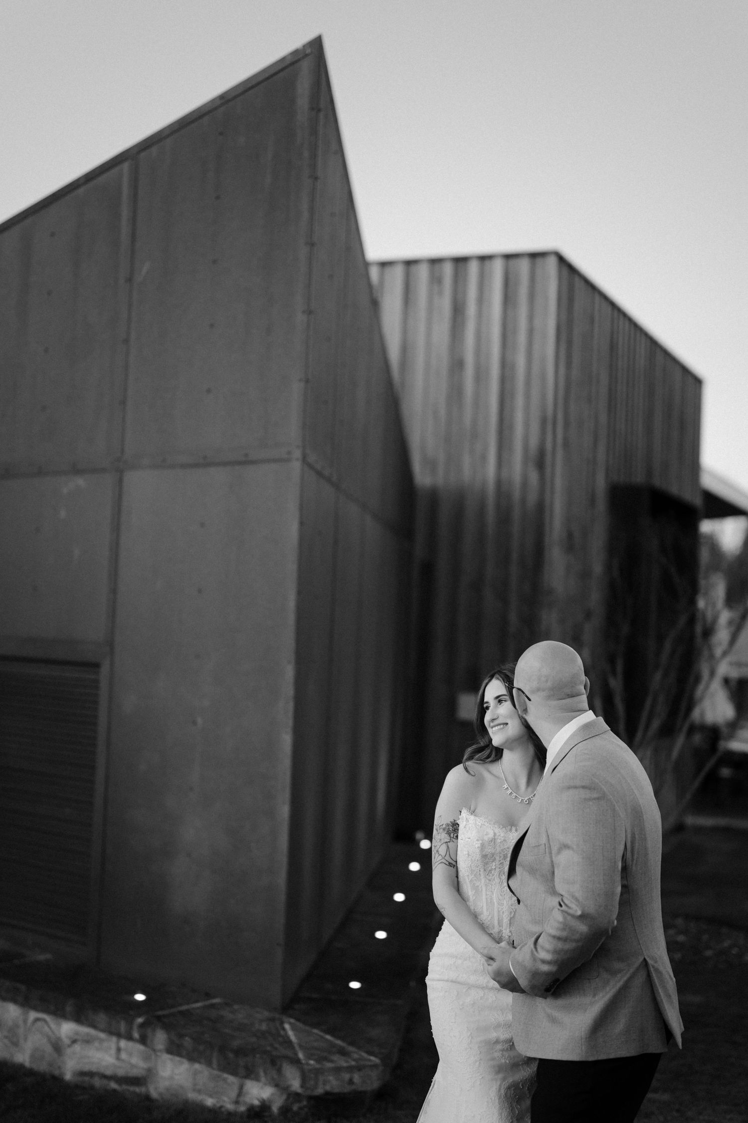 Black and white wedding photo of bride and groom embracing outdoors near modern architectural building at dusk.