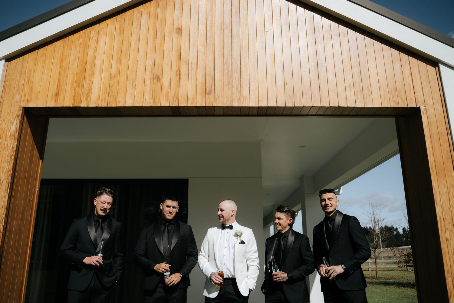 Groom in white tuxedo stands with four groomsmen in black suits outside a wooden barn venue.