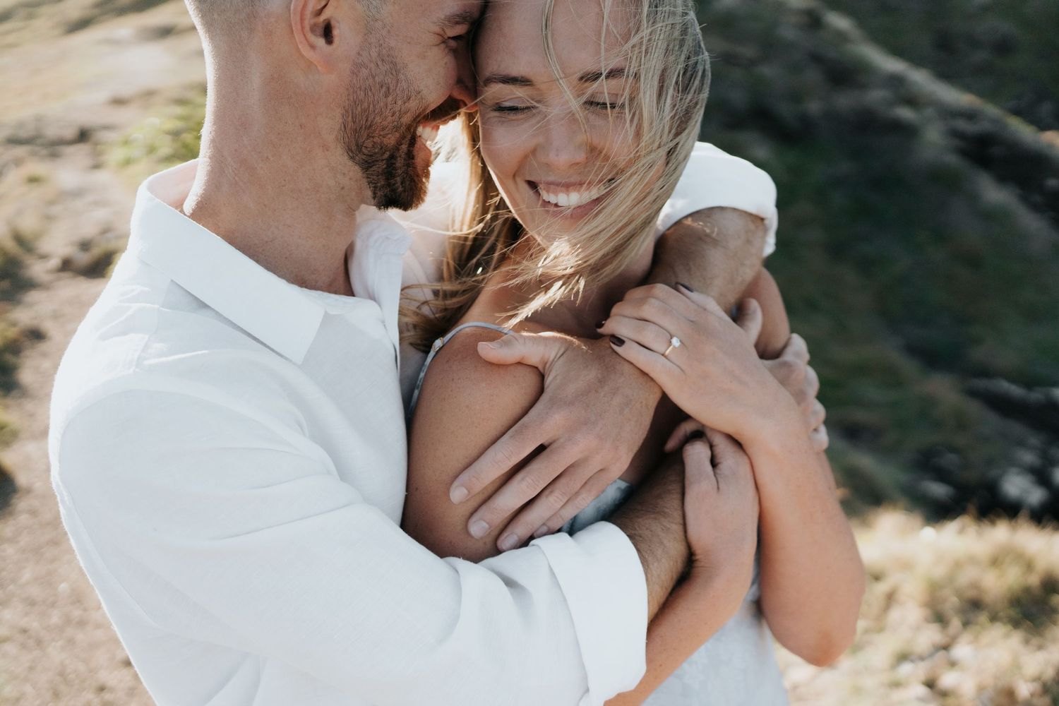 Happy couple embracing outdoors, woman wearing engagement ring, smiling joyfully in a romantic mountain setting.