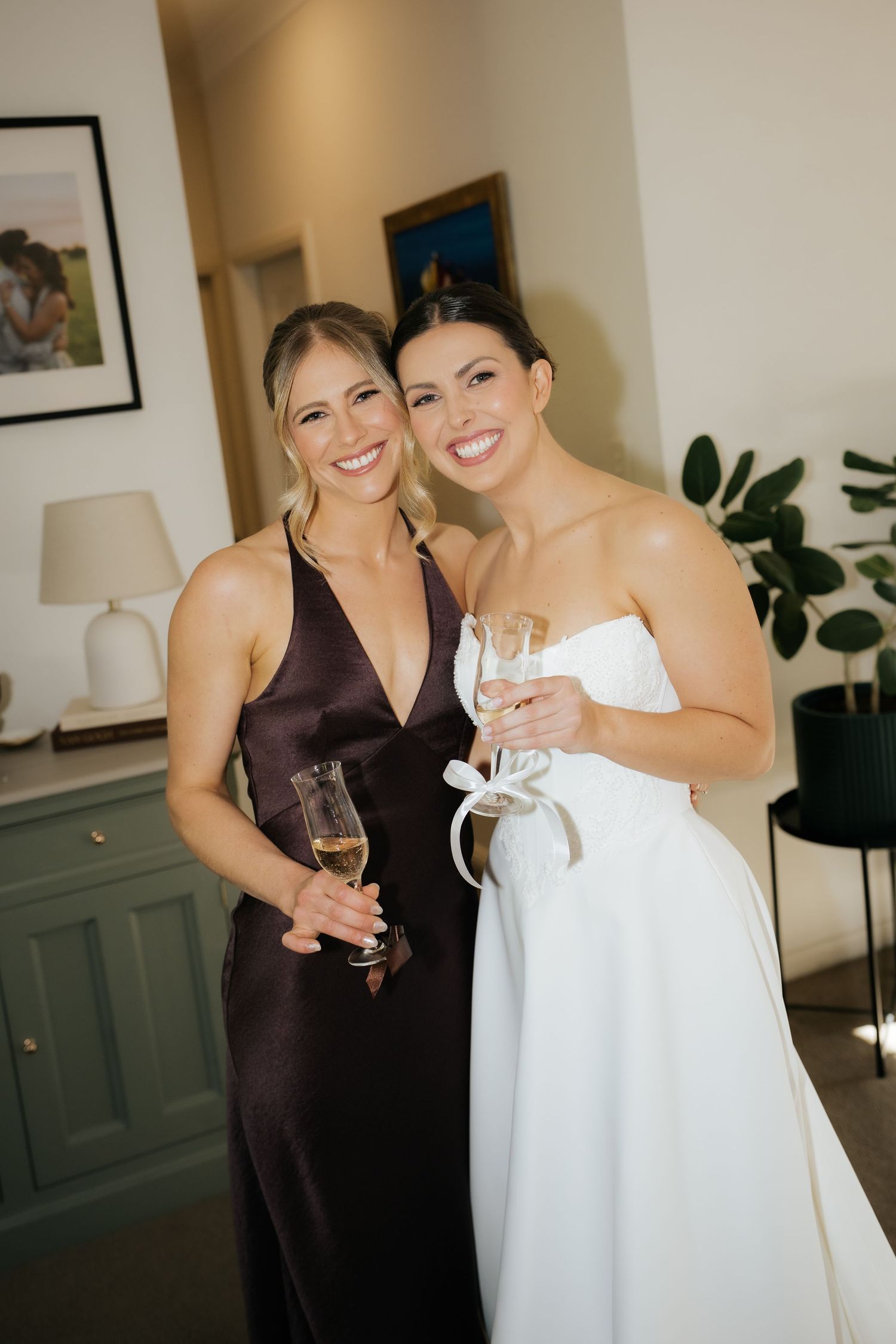 Two women smile holding champagne glasses, one in a white strapless wedding gown and one in a dark satin gown.
