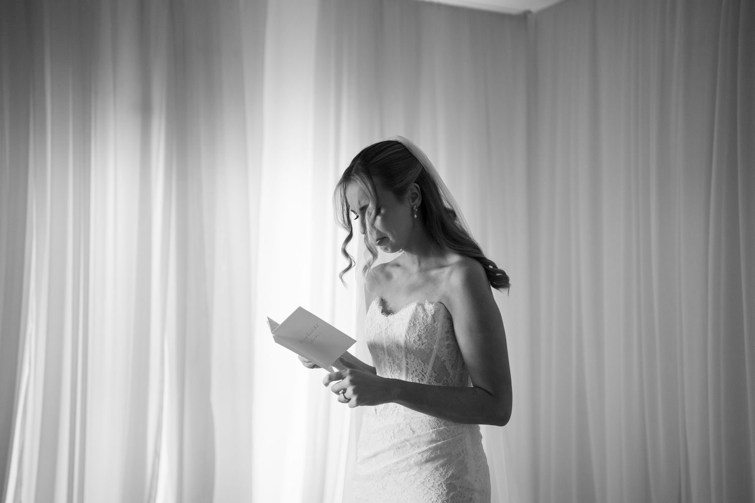 Bride in white gown reads wedding vows in black and white photo with soft curtain backdrop.
