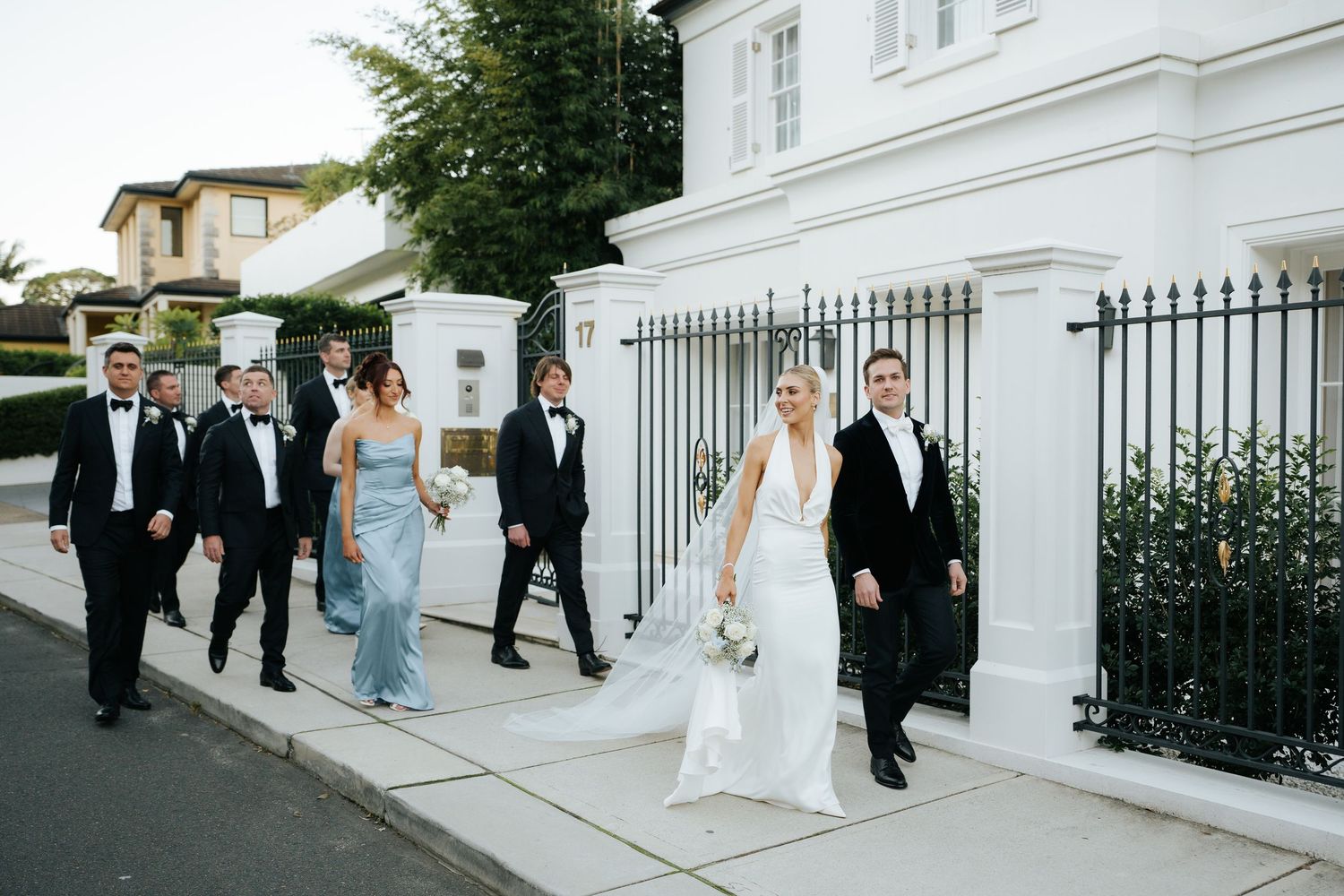 Wedding party in formal attire walking along a street past a white building with iron fence.