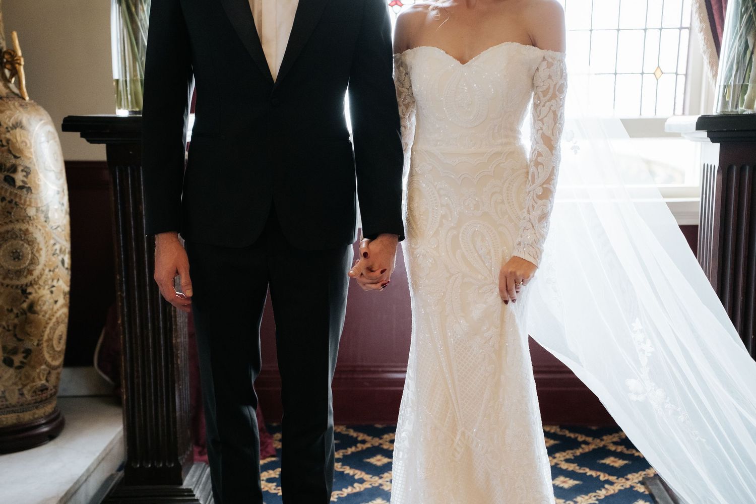 Bride in lace off-shoulder gown with veil and groom in black tuxedo holding hands at elegant wedding venue.
