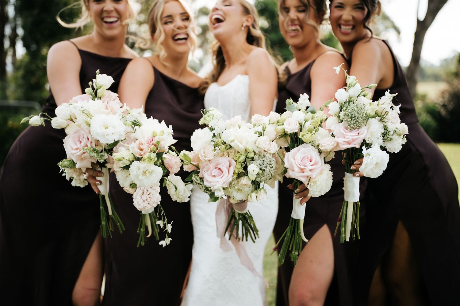 Bride in white gown with bridesmaids in dark dresses holding blush and white floral bouquets laughing outdoors.