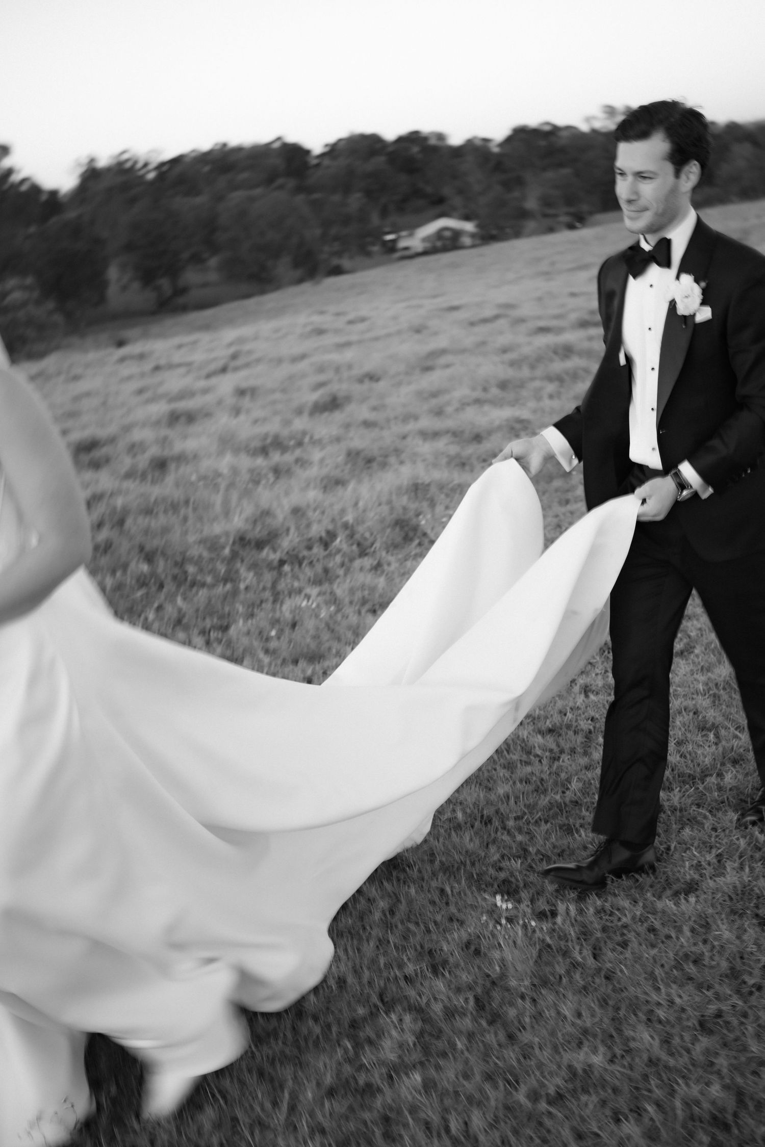 Black and white photo of groom in tuxedo holding bride's flowing wedding dress train in open field.
