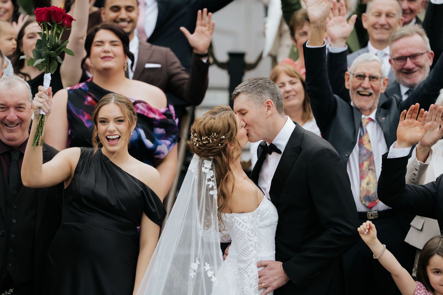 Bride and groom share a kiss surrounded by cheering wedding guests, with a bridesmaid in black holding red roses.