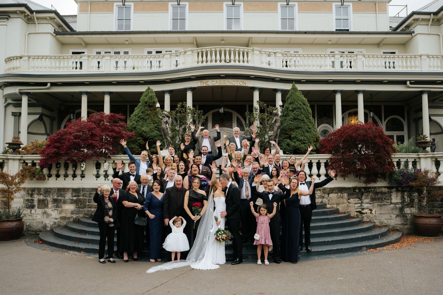 Large wedding party celebrating on grand mansion steps with bride, groom, and guests cheering outdoors.