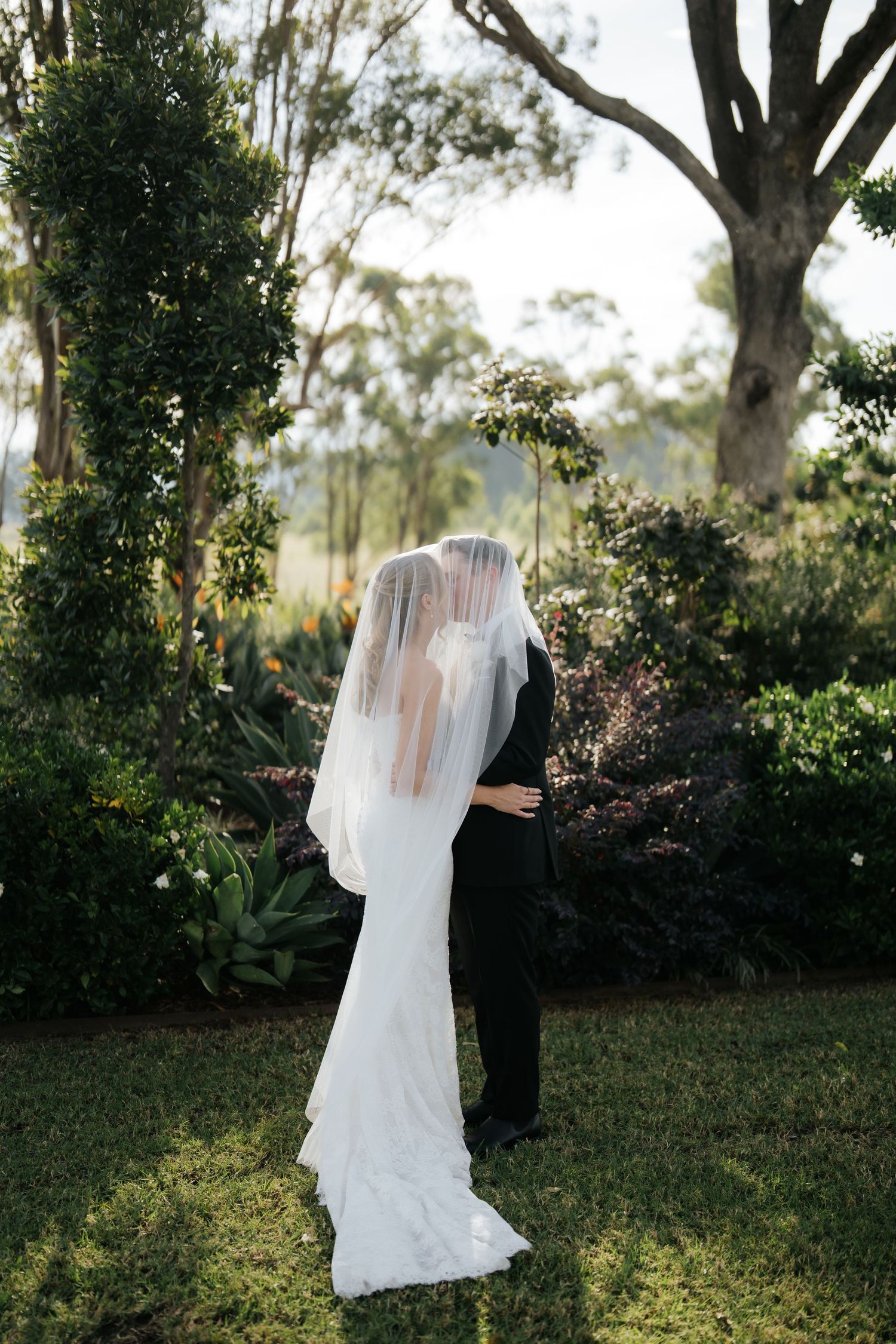 Bride and groom share a kiss under a flowing veil in a lush garden setting on their wedding day.