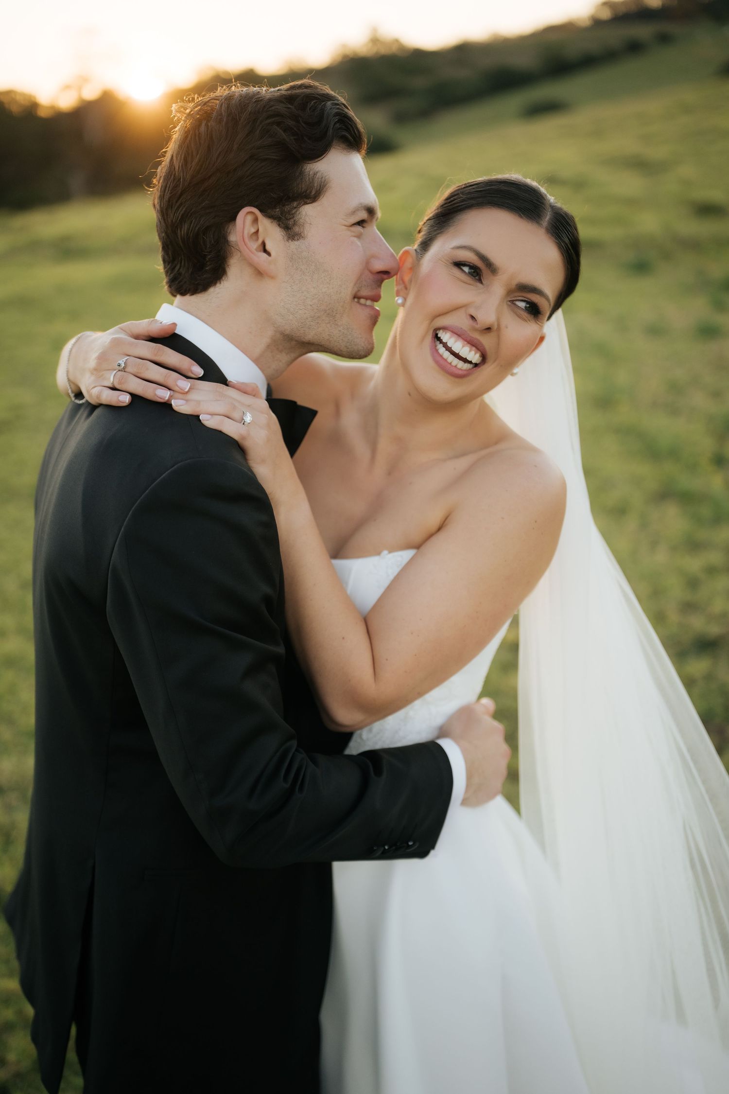 Smiling bride in strapless gown and veil embracing groom in black tuxedo during golden hour sunset.
