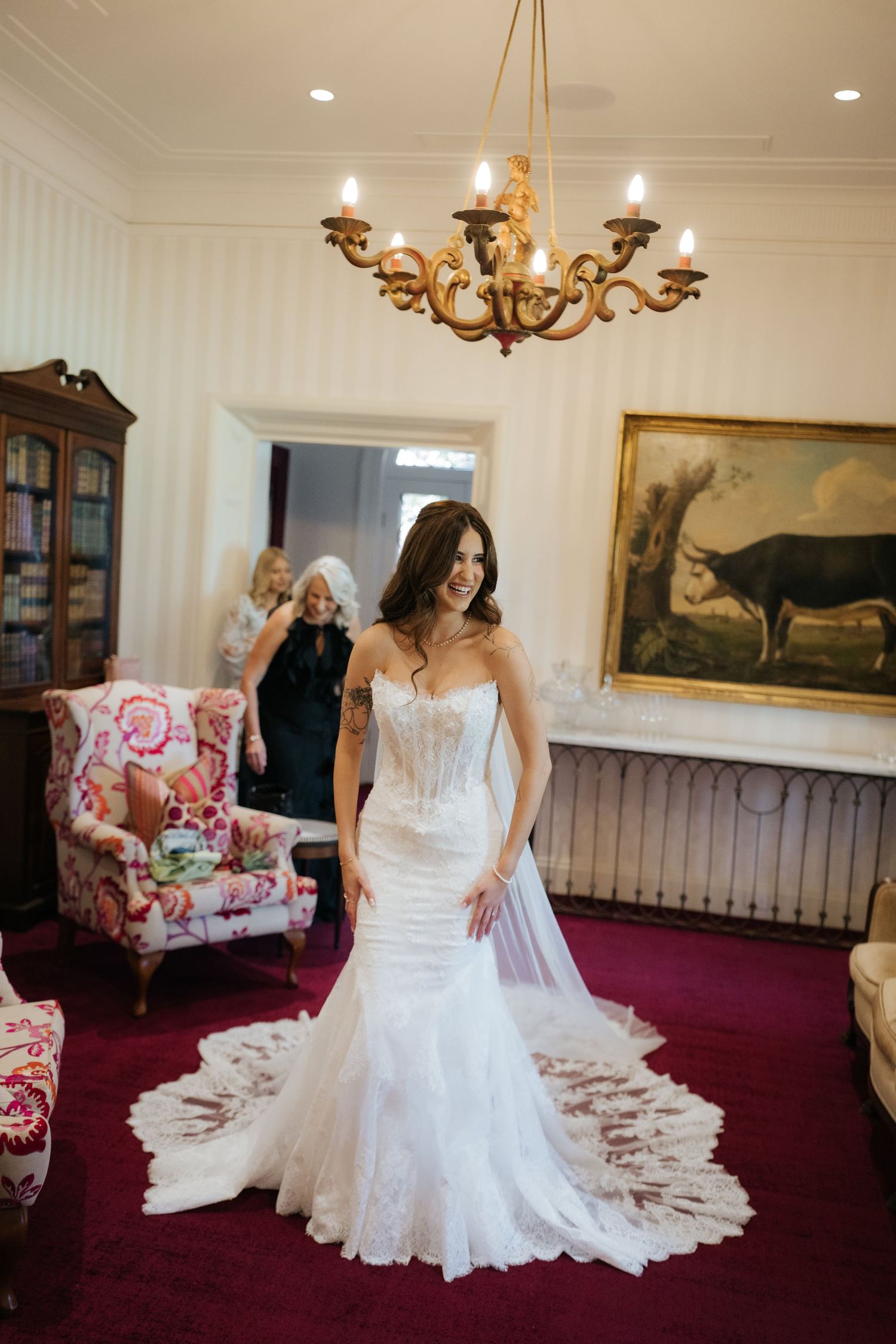 Bride in a strapless lace mermaid wedding gown stands under a chandelier in an elegant room with a painting.